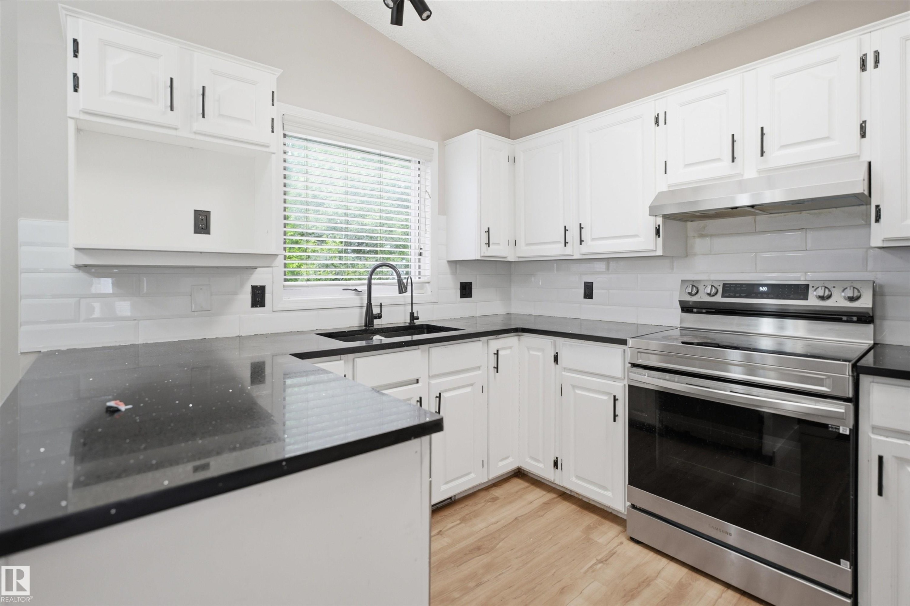 Kitchen featuring white cabinetry with dark hardware, dark countertops, white subway tile backsplash, stainless steel range with overhead hood, and light wood-finish flooring - 3220 42A Avenue, Edmonton, AB