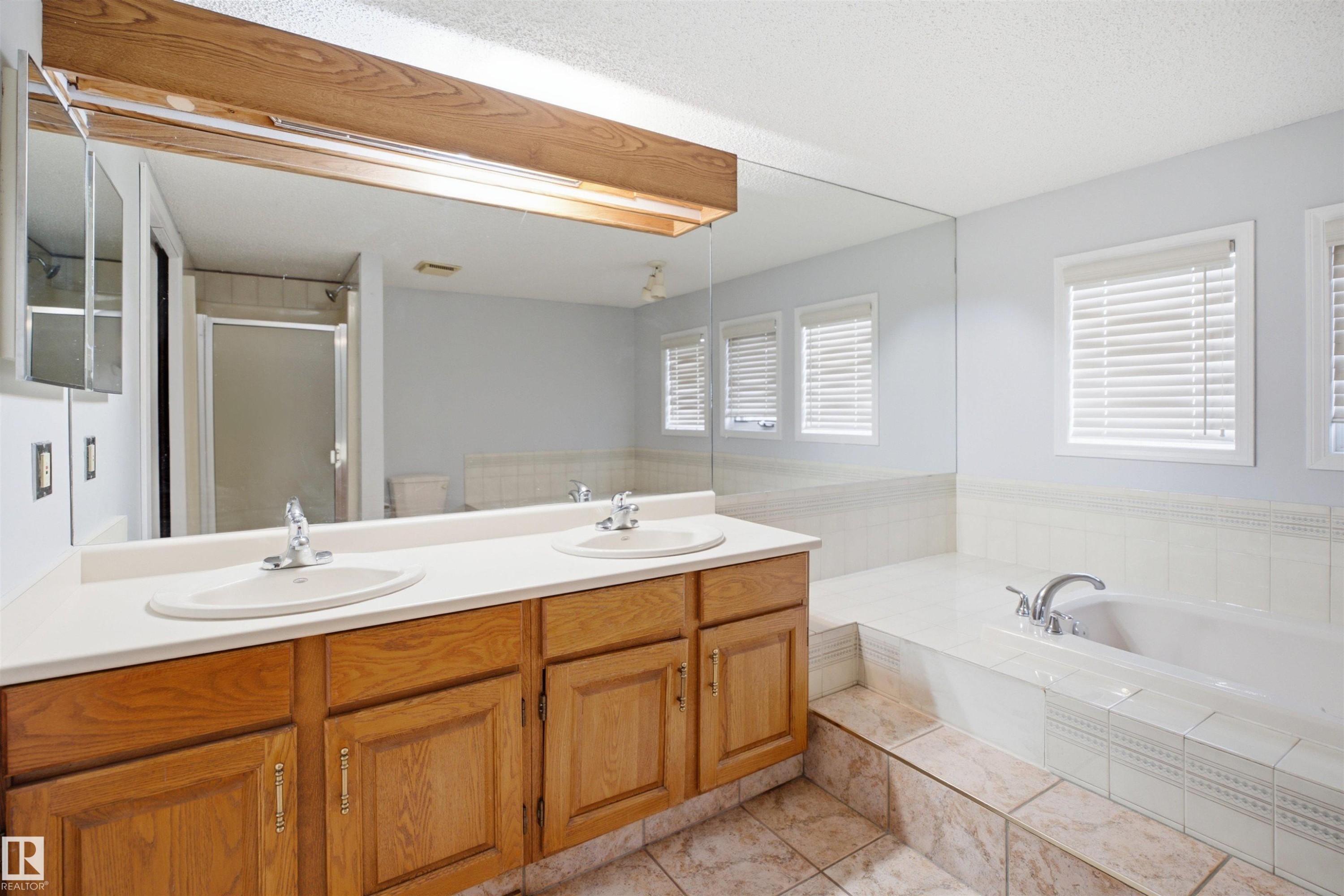 Bathroom featuring a dual vanity with wood cabinetry, a large built-in mirror, a tiled soaking tub, and multiple windows with blinds - 3220 42A Avenue, Edmonton, AB
