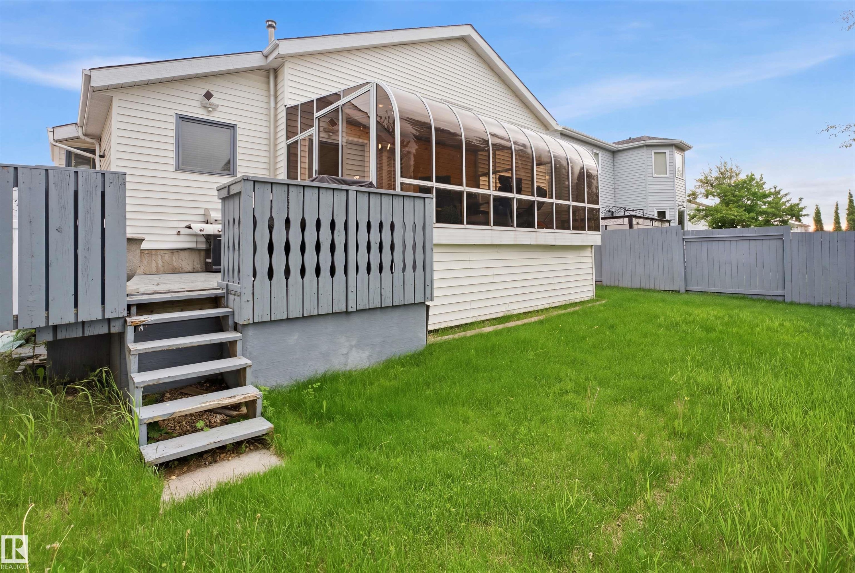 Rear exterior featuring a sunroom addition with curved glass panels, white siding, a multi-level deck with gray railings, steps leading to a grassy yard, and a gray privacy fence - 3220 42A Avenue, Edmonton, AB