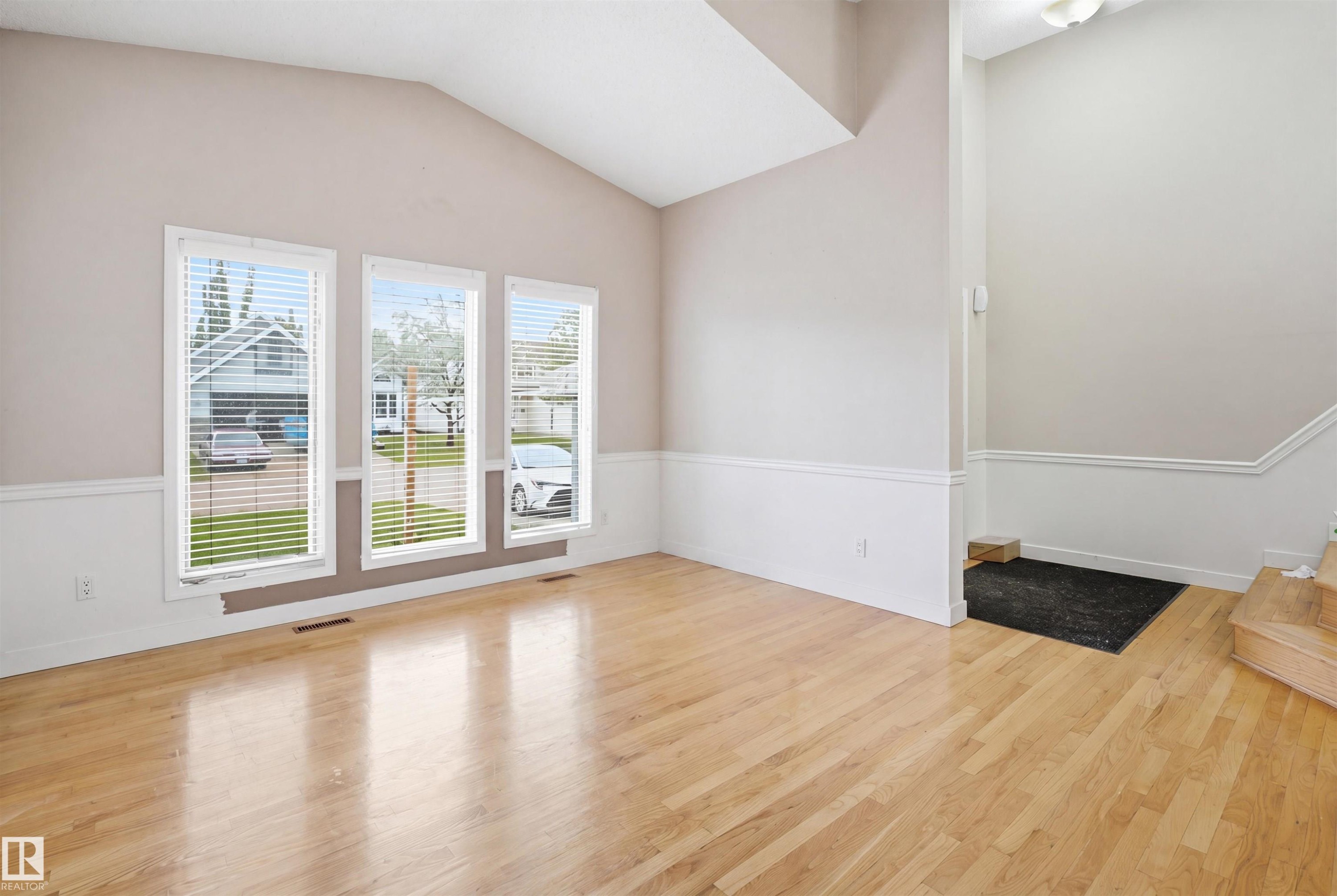 Bright interior featuring wood-finish flooring, three vertical windows with blinds, a vaulted ceiling, and white wainscoting - 3220 42A Avenue, Edmonton, AB