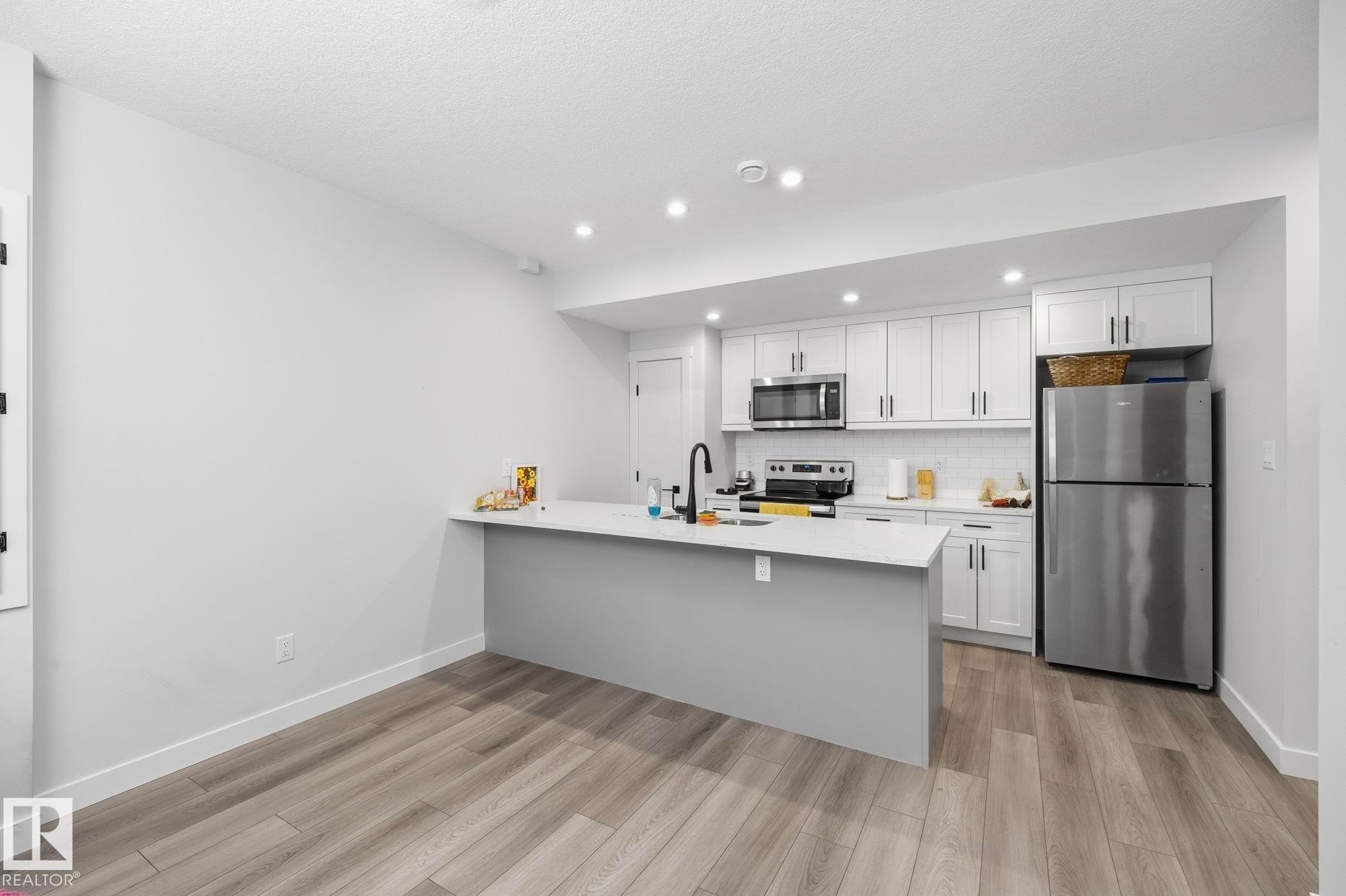 Contemporary kitchen featuring stainless steel appliances, white cabinetry, a white subway tile backsplash, a center island with a sink, and wood-finish flooring - 11012 132 Street, Edmonton, AB