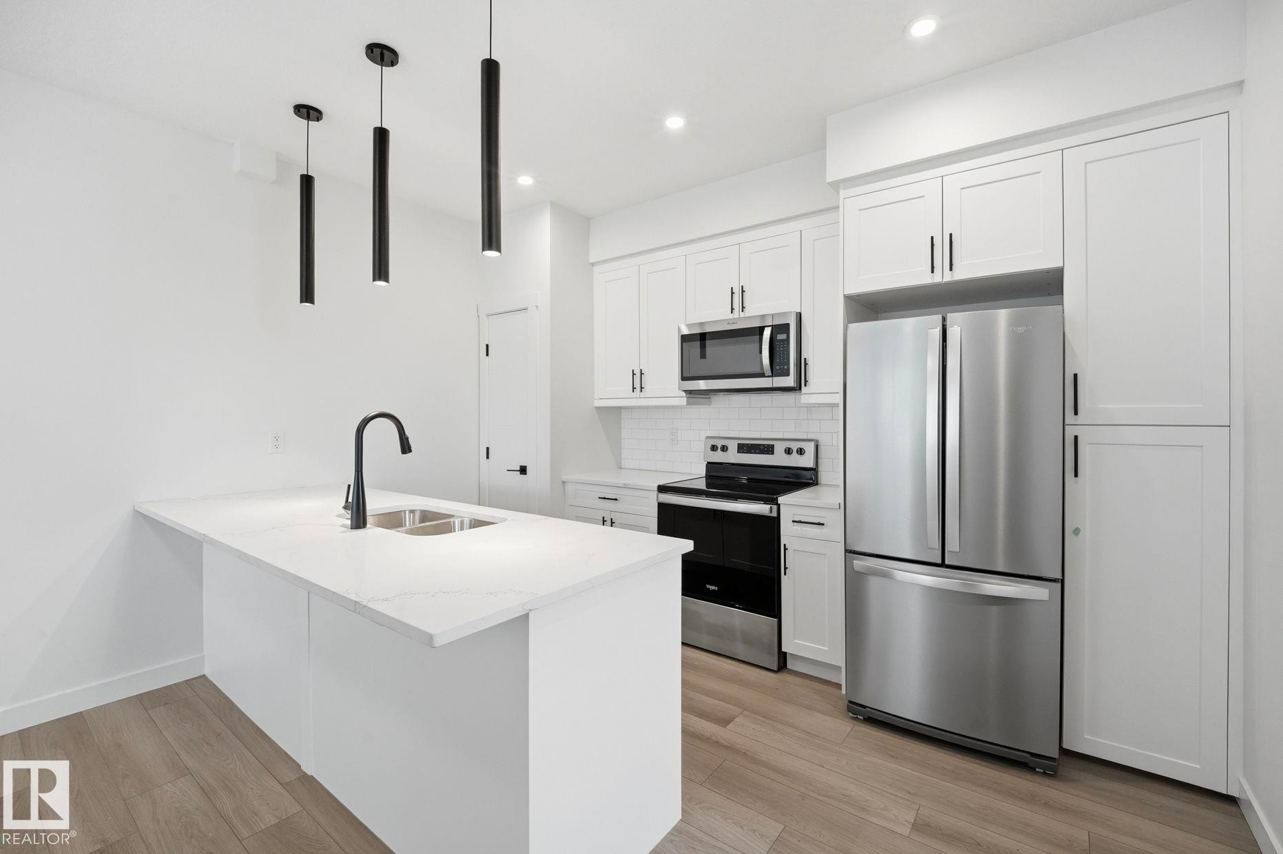 Kitchen featuring a white island with an undermount double sink and matte black faucet, pendant lighting, wood-finish flooring, stainless steel appliances, and white shaker cabinetry - 11012 132 Street, Edmonton, AB