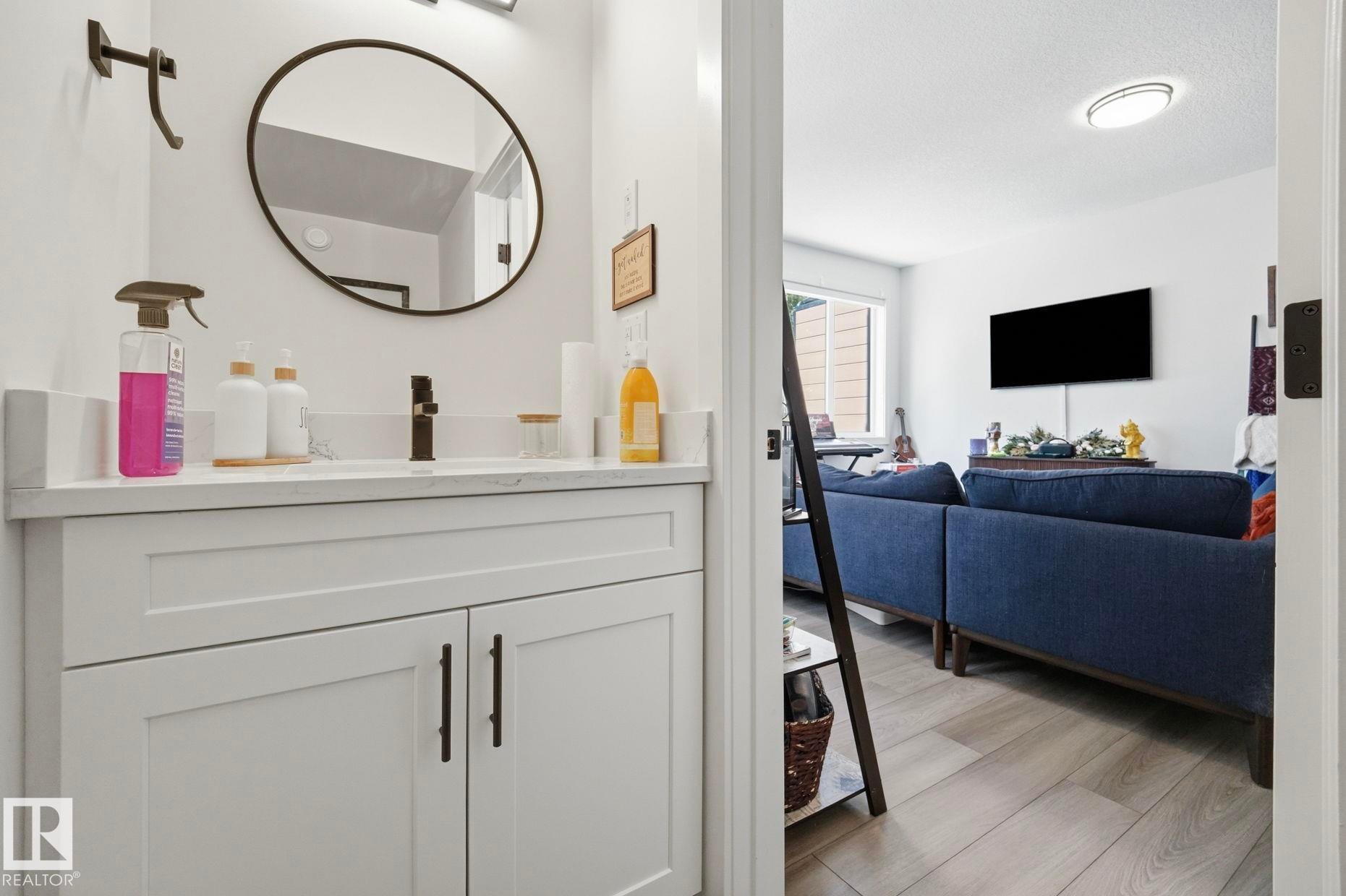 Modern bathroom vanity featuring white shaker-style cabinetry with matte black hardware, a pristine white countertop, and a circular black-framed mirror - 11007 132 Street, Edmonton, AB