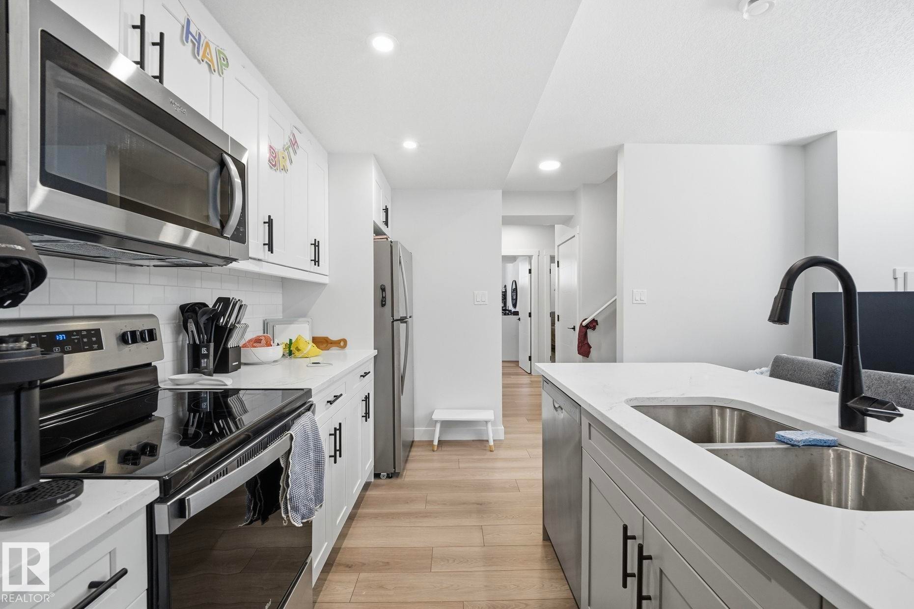 Contemporary kitchen featuring stainless steel appliances, white subway tile backsplash, light wood-finish flooring, and an island with an undermount double-basin sink and matte black faucet - 11007 132 Street, Edmonton, AB