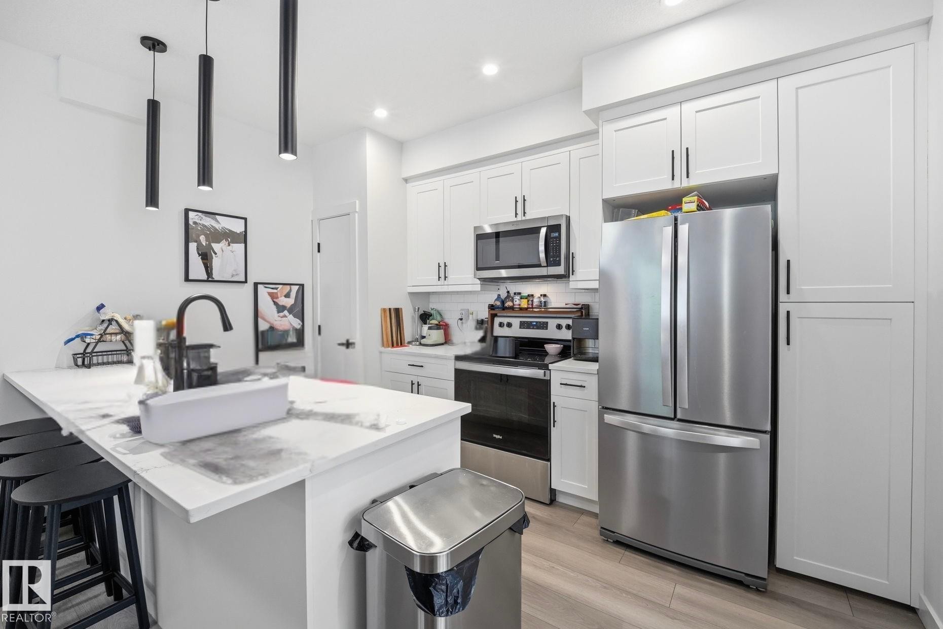 Modern kitchen featuring white shaker cabinetry, stainless steel appliances, a center island with a white countertop, and dark pendant lighting - 11007 132 Street, Edmonton, AB