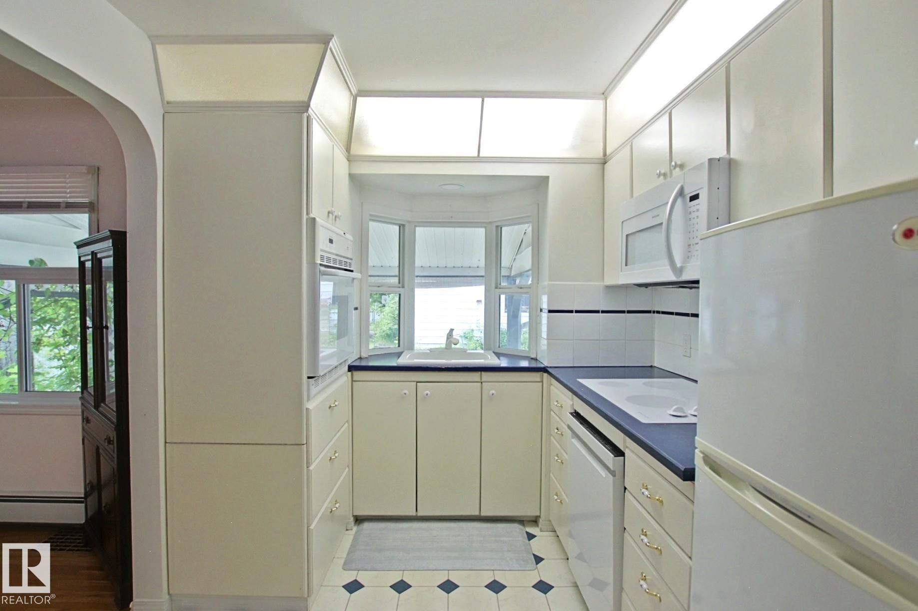 Galley kitchen featuring a bay window, white cabinetry, dark countertops, built-in microwave, and tile flooring with a geometric pattern - 11242 56 Street, Edmonton, AB