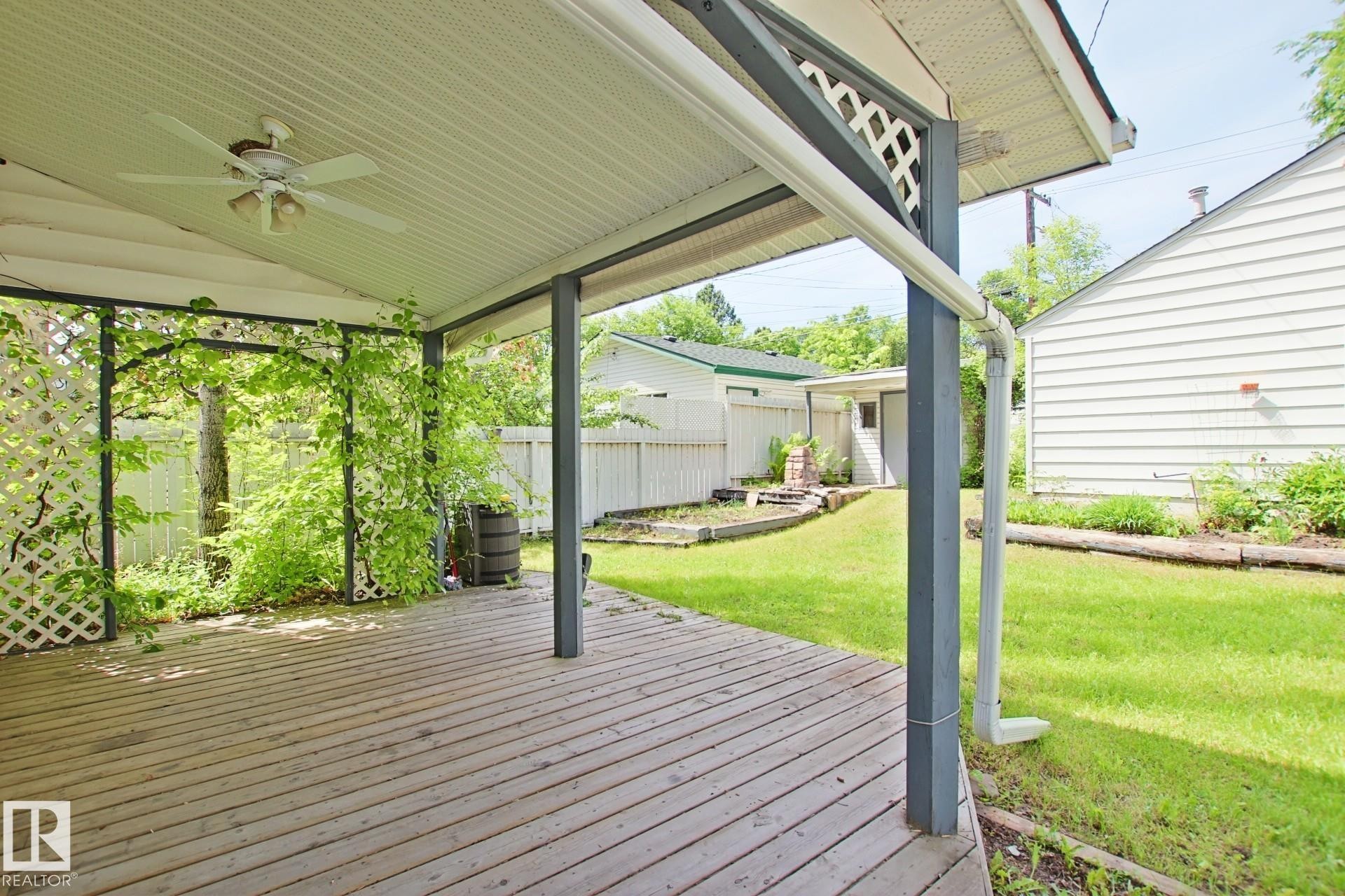 Covered outdoor deck with a white ceiling fan and integrated lighting, featuring a wood-plank surface and white lattice accents with climbing greenery - 11242 56 Street, Edmonton, AB