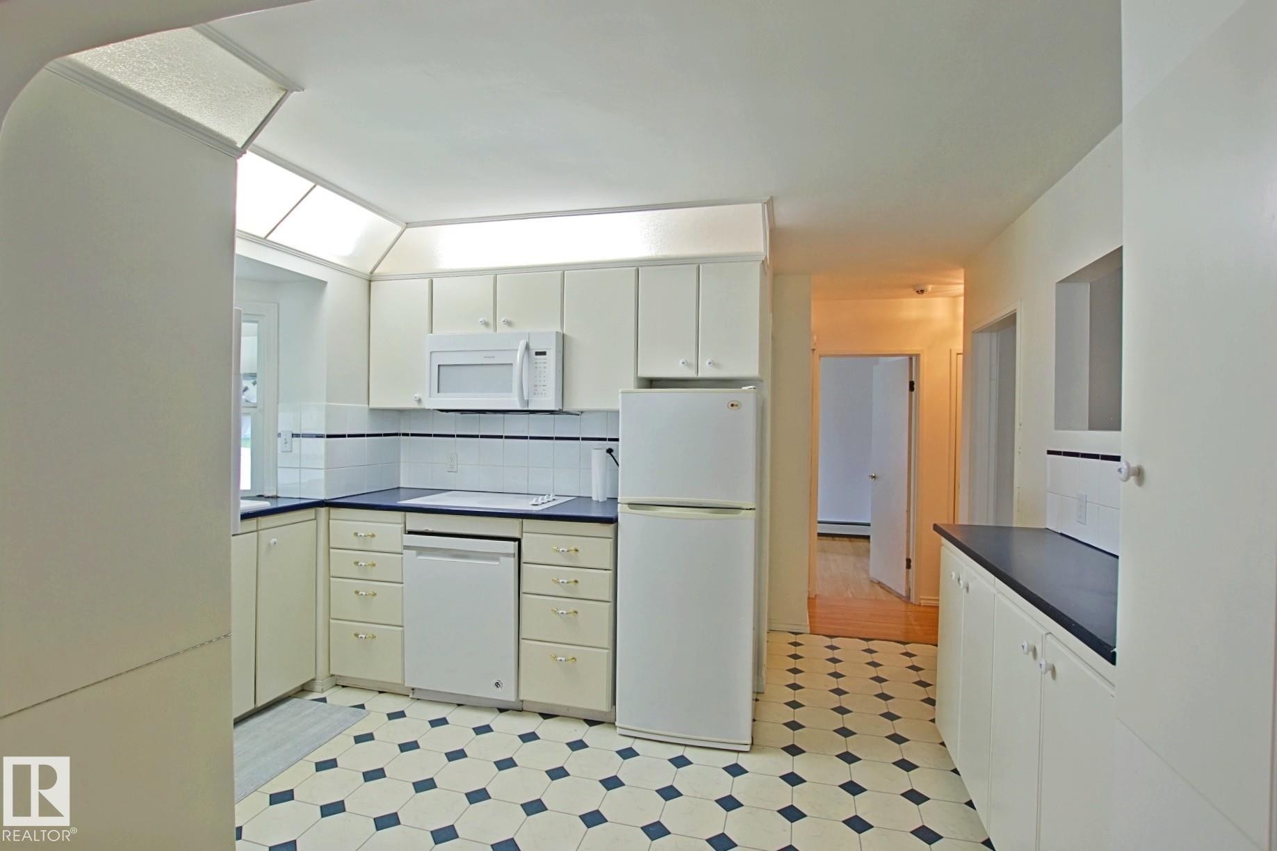 Kitchen featuring white cabinetry, integrated white appliances, dark countertops, a white tile backsplash with a dark border, and patterned hexagonal floor tiles - 11242 56 Street, Edmonton, AB