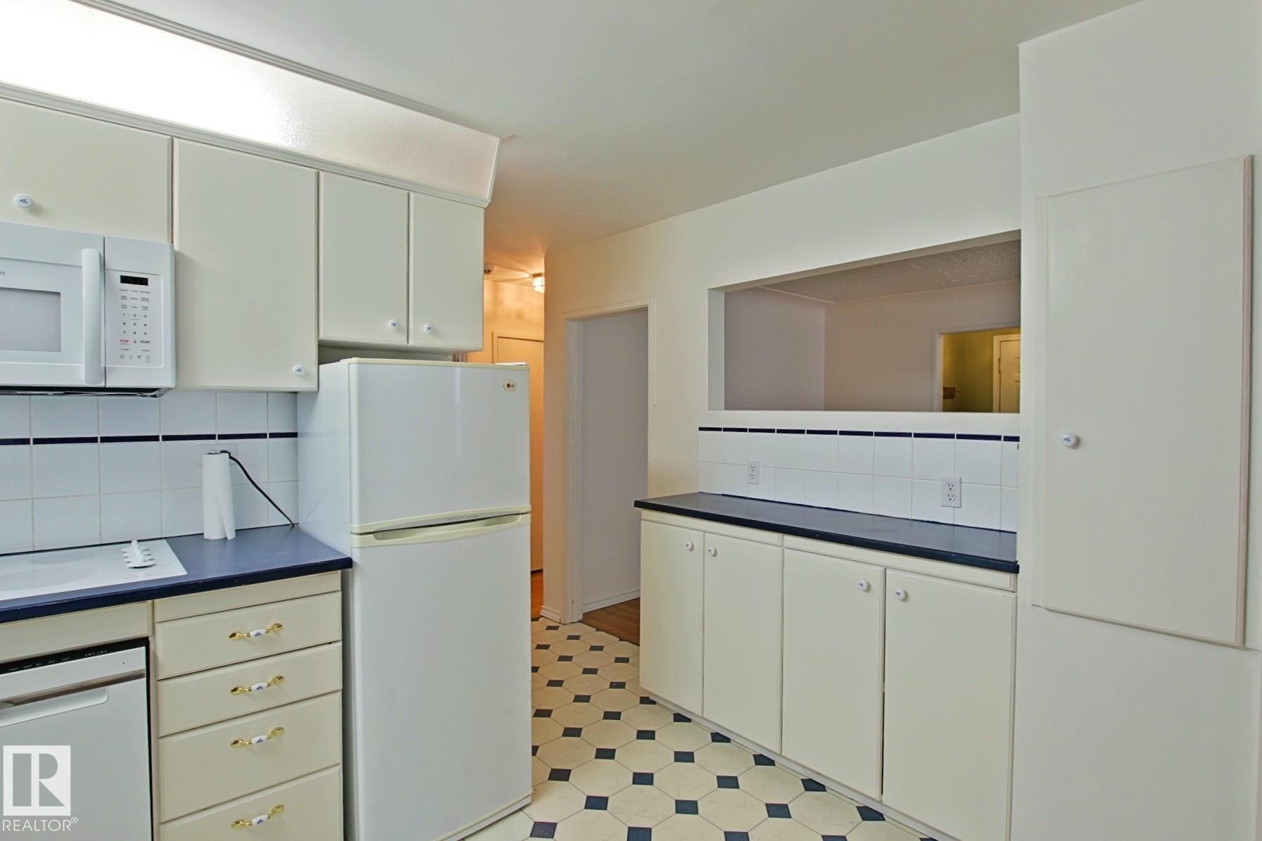 Kitchen featuring white cabinetry, dark countertops, a white tile backsplash with a dark accent line, and patterned floor tile - 11242 56 Street, Edmonton, AB