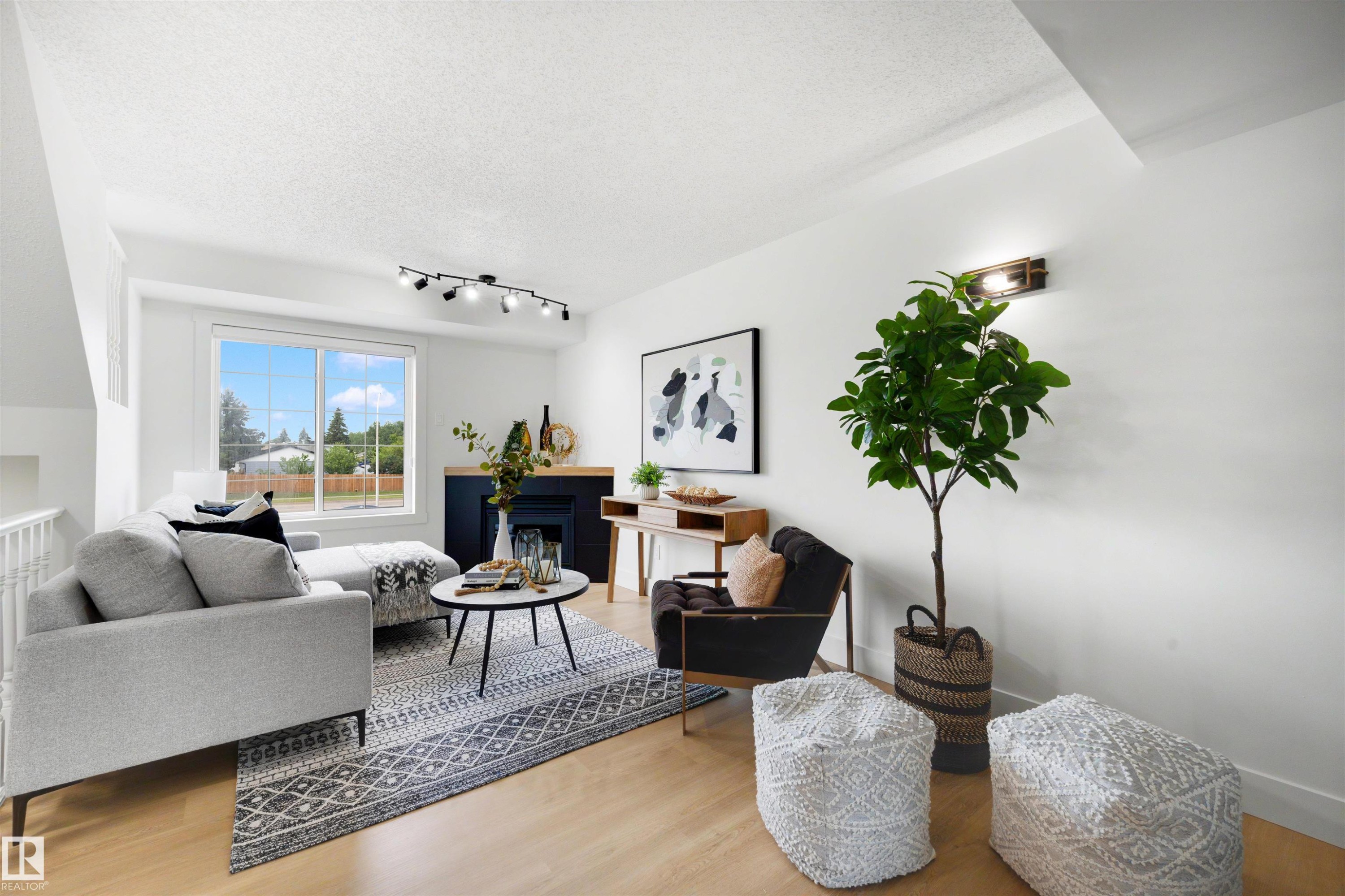 Bright living area featuring wood-finish flooring, a large window, a fireplace with a dark surround, track lighting, and a wall-mounted fixture - 12611 47 Street, Edmonton, AB