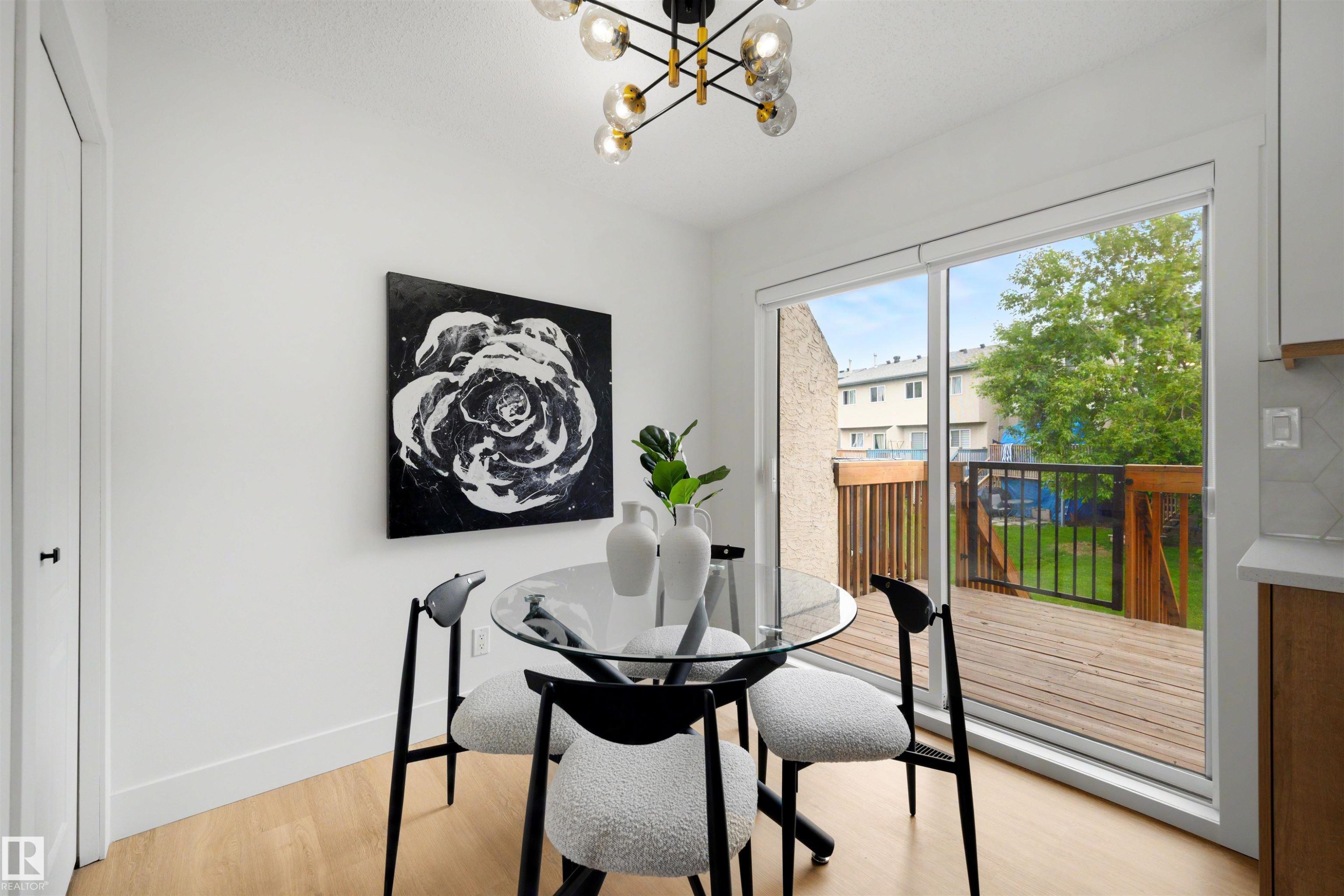 Bright dining area featuring light wood-finish flooring, a modern chandelier with spherical glass shades, and a sliding glass door providing direct access to an outdoor wooden deck with black metal railings - 12611 47 Street, Edmonton, AB
