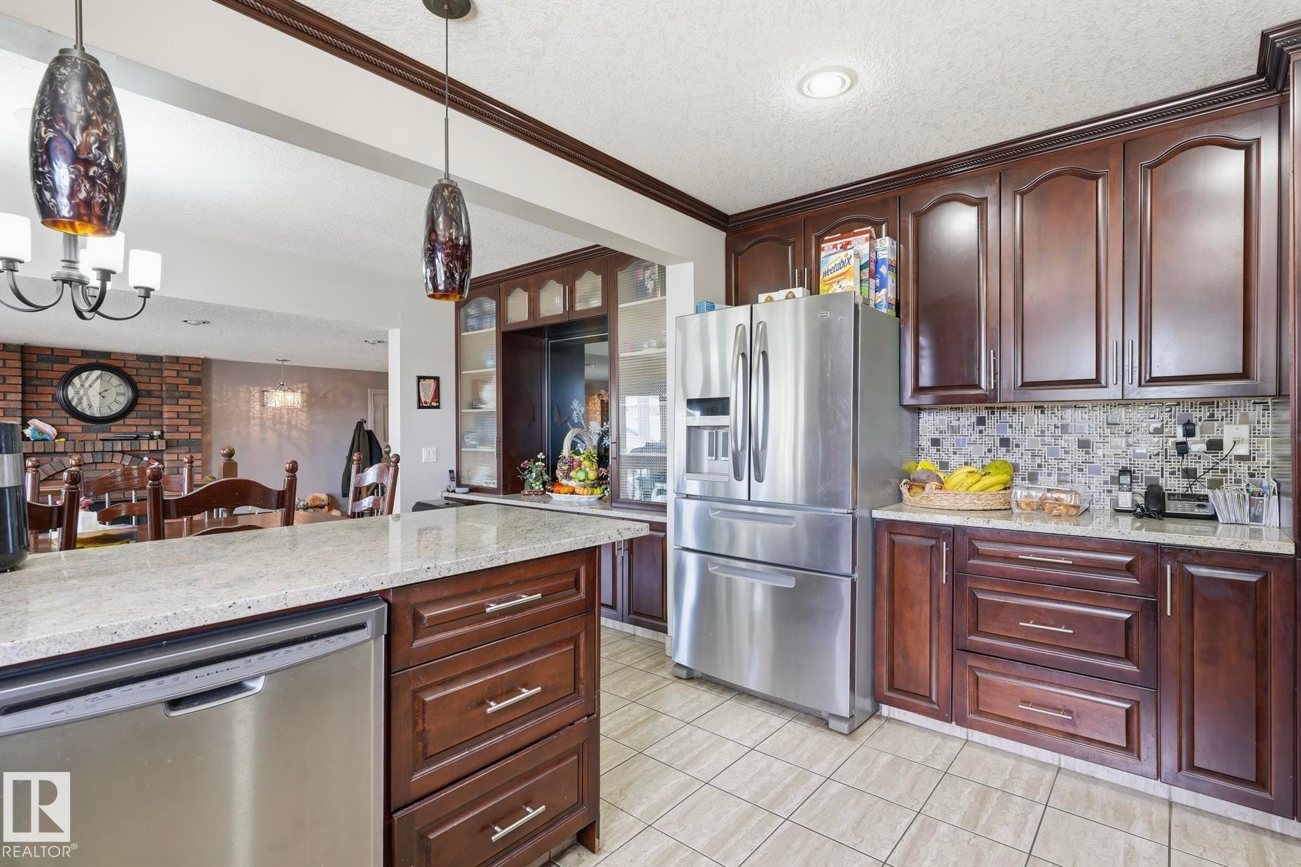 Kitchen featuring rich wood cabinetry with crown molding, stainless steel appliances, granite-look countertops, and a mosaic tile backsplash - 12131 101 Street Nw, Edmonton, AB