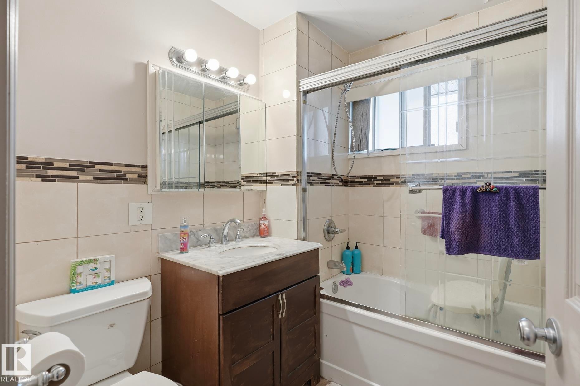 Tiled bathroom featuring a combination tub and shower with glass enclosure, a window, and a vanity with a white countertop - 12131 101 Street Nw, Edmonton, AB