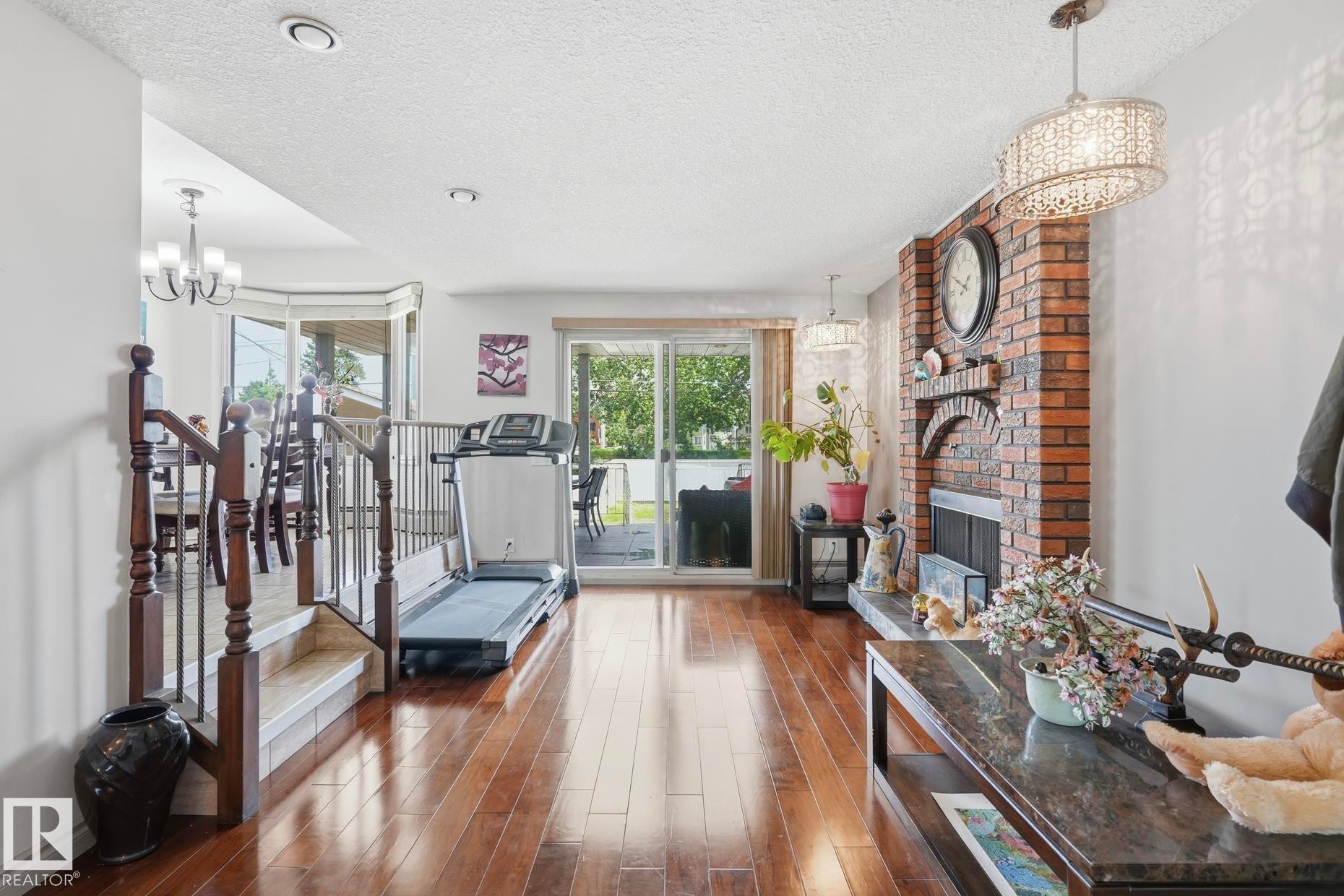 Spacious living area featuring wood-finish flooring, a brick fireplace with an arched opening, and a contemporary chandelier - 12131 101 Street Nw, Edmonton, AB