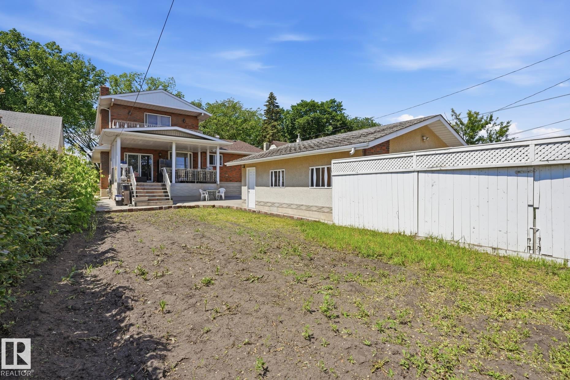 Expansive backyard space with a brick exterior home featuring a covered patio, second-story balcony, and a separate structure with a white privacy fence - 12131 101 Street Nw, Edmonton, AB