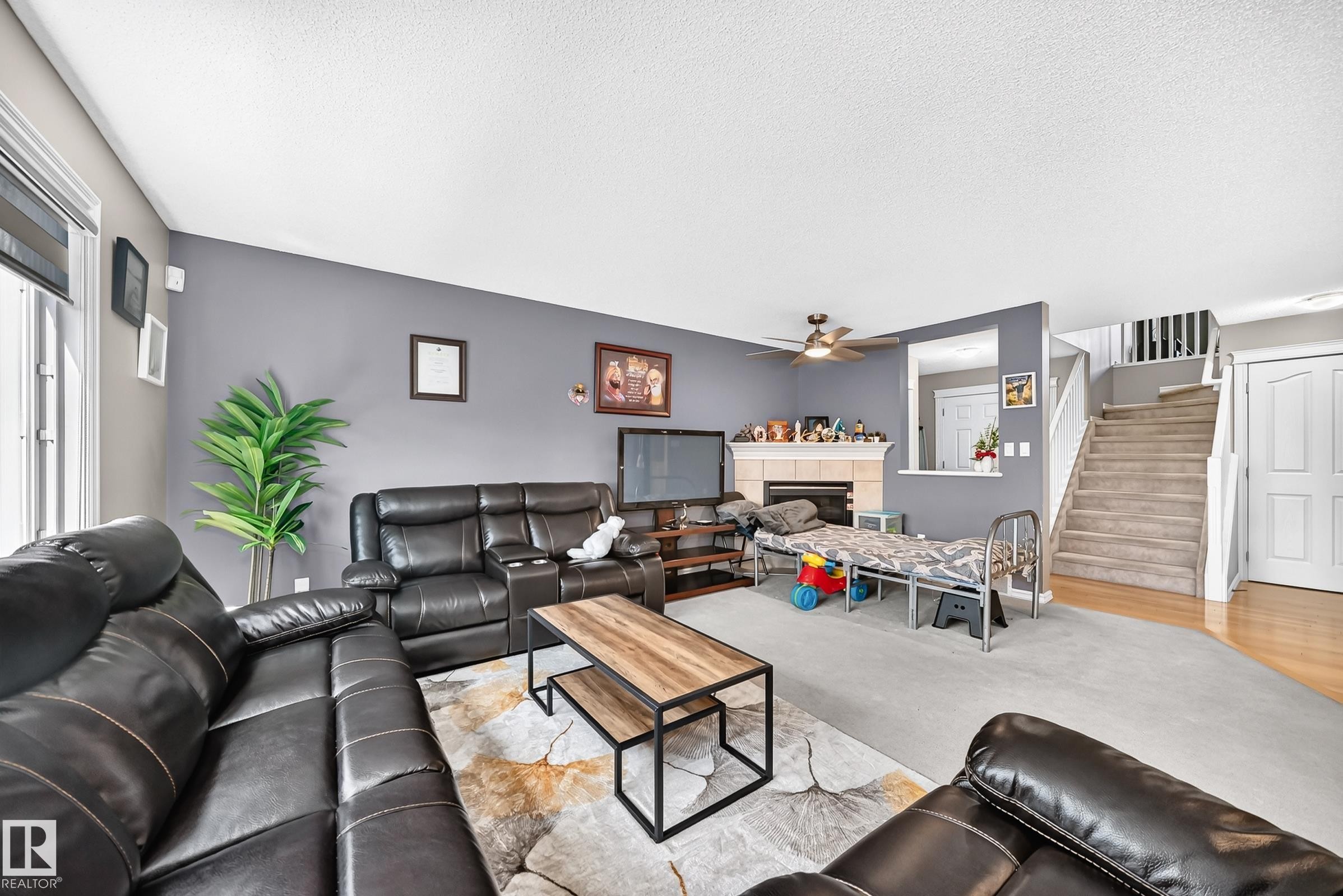 Spacious living area featuring a central fireplace with mantle, a ceiling fan, a staircase with carpeting, and wood-finish flooring extending into the hallway - 3224 26 Street, Edmonton, AB