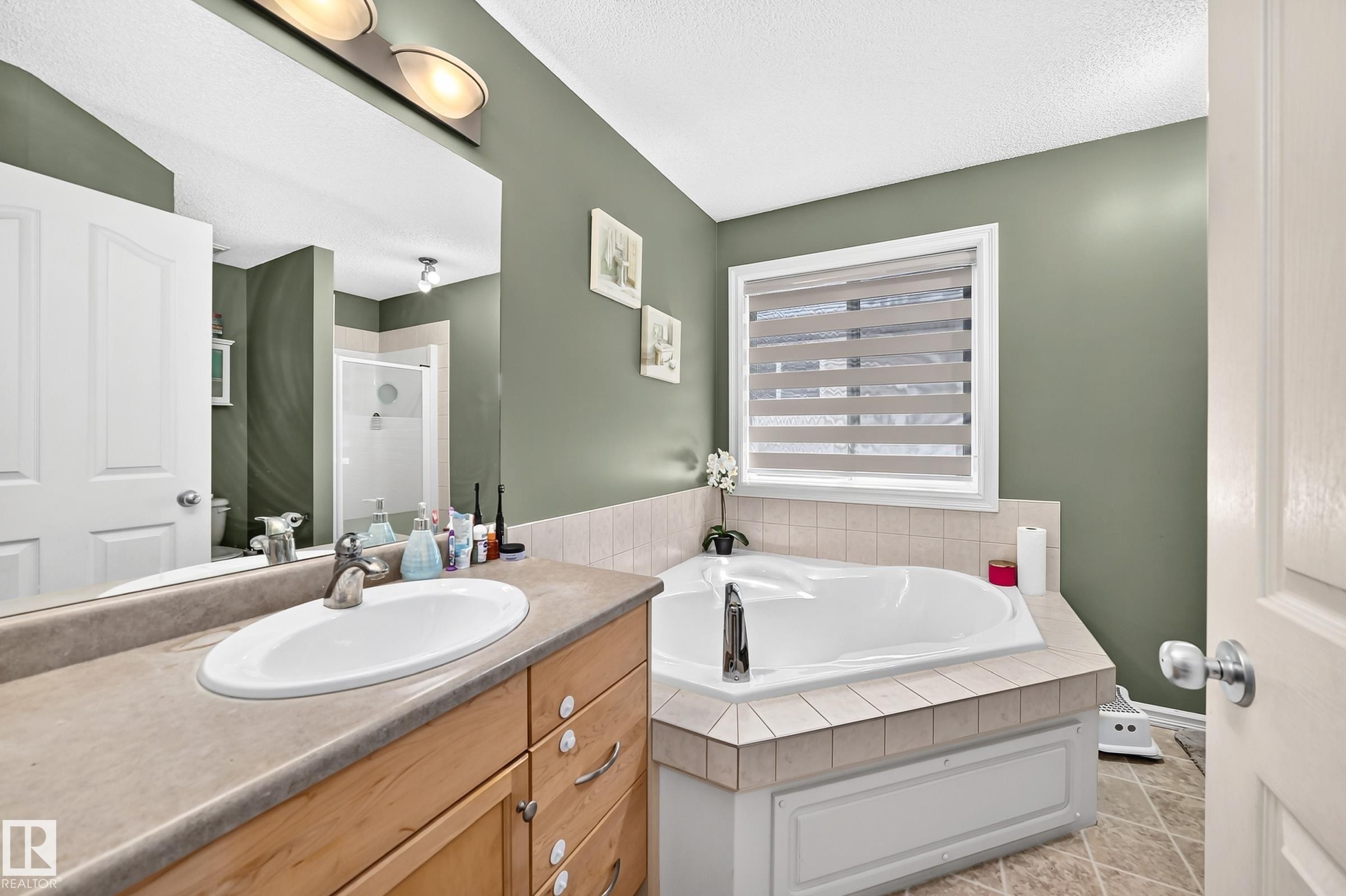 Bathroom featuring an integrated jetted tub with tile surround, a single vanity with a laminate countertop and undermount sink, and a mirrored wall with an overhead light fixture - 3224 26 Street, Edmonton, AB