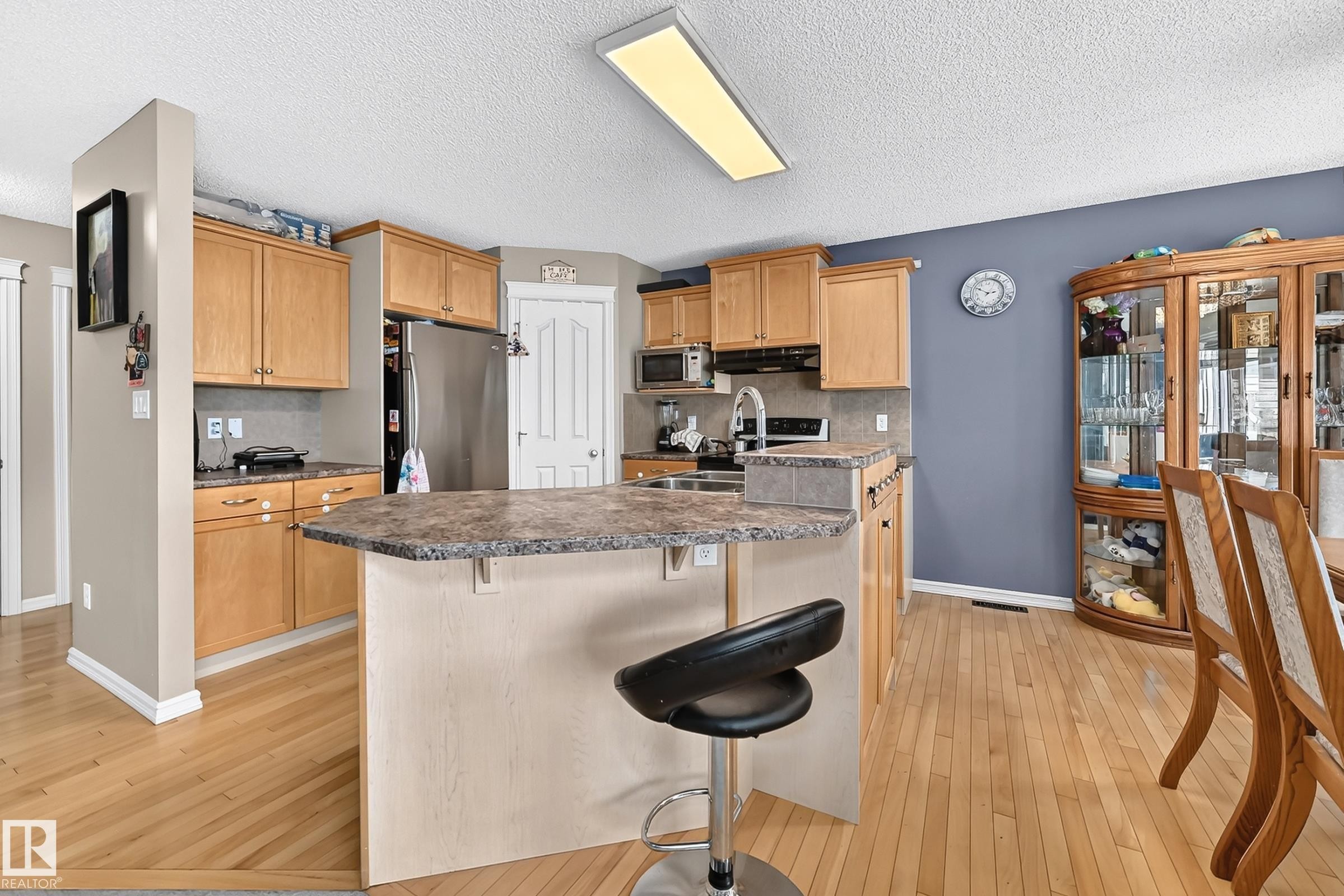 Kitchen featuring wood-finish flooring, light wood cabinetry, stone-look countertops, stainless steel appliances, and a central island with breakfast bar seating - 3224 26 Street, Edmonton, AB