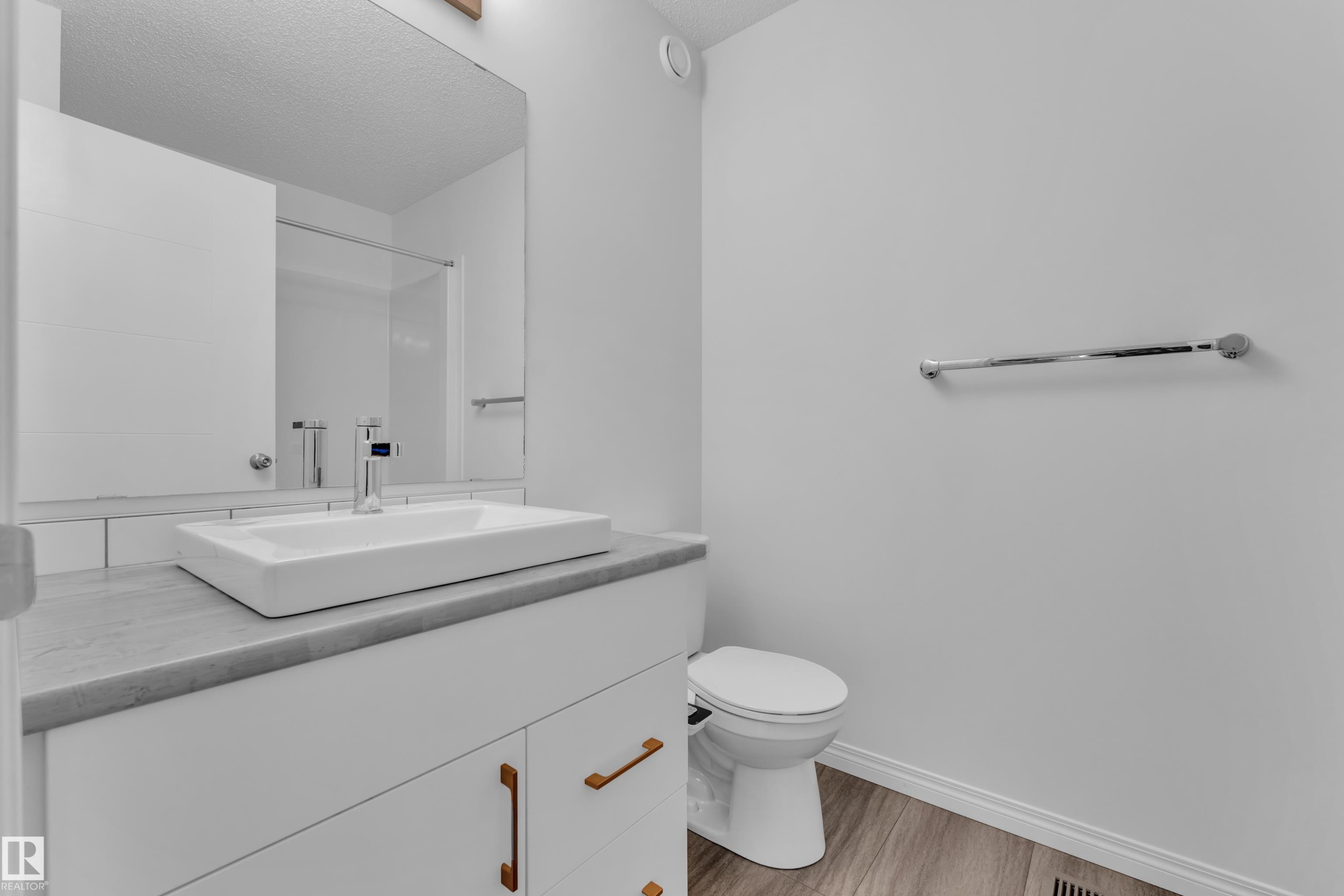 Modern bathroom featuring a white vessel sink on a gray countertop, white cabinetry with copper-toned hardware, a wall-mounted mirror, and wood-finish flooring - 144 Sunwapta Way, Edmonton, AB
