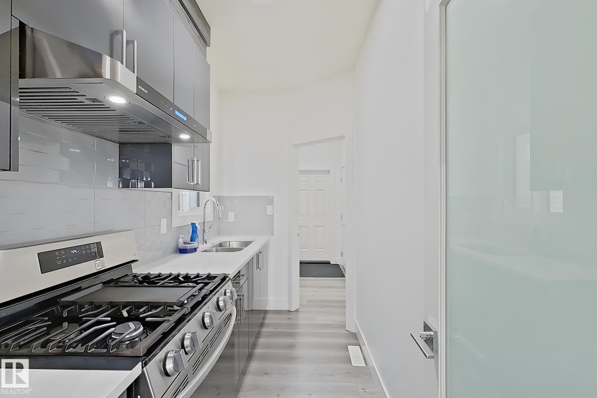 Modern kitchen featuring stainless steel appliances, a gas range with an overhead vent hood, white countertops, and gray wood-finish flooring - 415 42 Street, Edmonton, AB