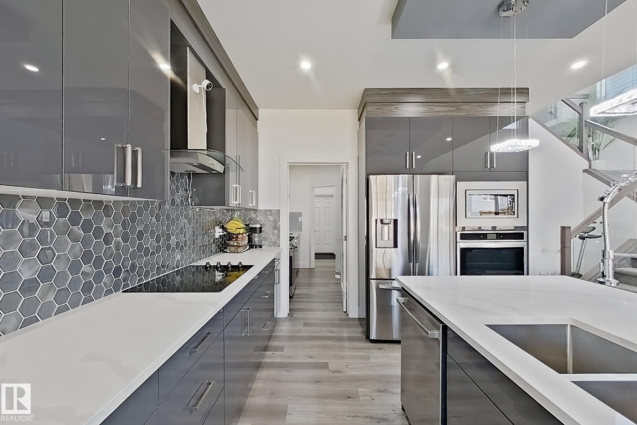 Contemporary kitchen featuring sleek gray cabinetry, light countertops, integrated stainless steel appliances, a hexagonal tile backsplash, and wood-finish flooring - 415 42 Street, Edmonton, AB