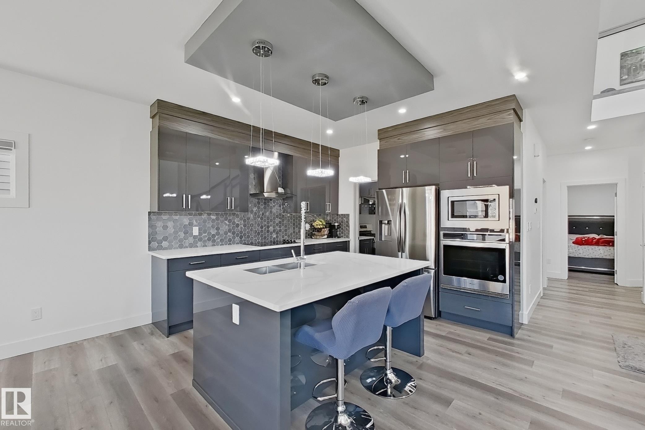 Contemporary kitchen featuring an island with a white countertop and undermount sink, grey high-gloss cabinetry, stainless steel appliances, and a grey hexagon tile backsplash - 415 42 Street, Edmonton, AB