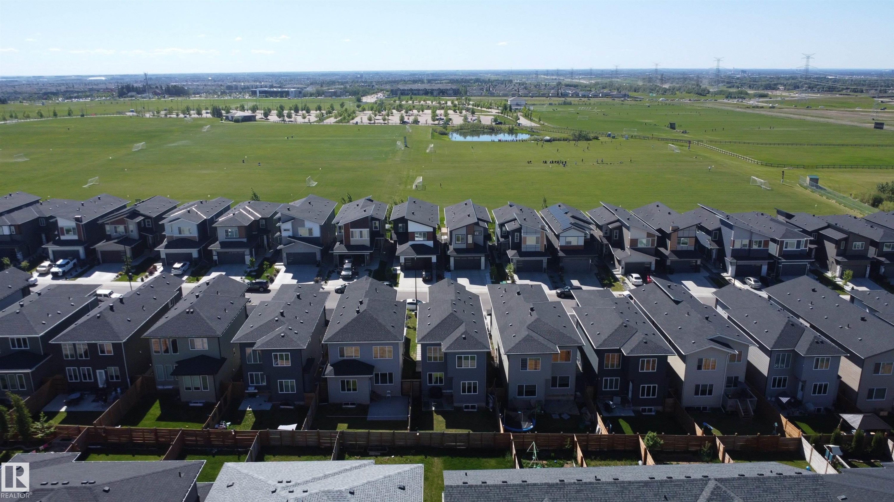 Aerial perspective showcasing a collection of modern residential properties with dark shingle roofs, individual private yards, and direct access to a large green field with a distant pond - 415 42 Street, Edmonton, AB