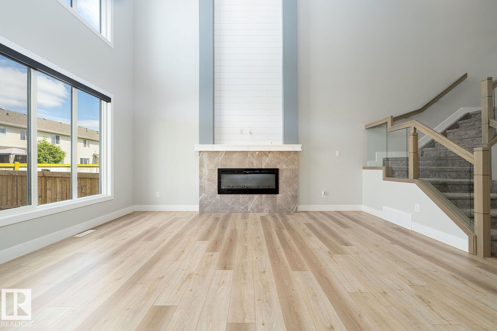 Two-story living area featuring a light wood-finish flooring, a contemporary fireplace with a stone-finish surround, high ceilings, large windows, and a staircase with glass panel railings - 17320 9 Avenue, Edmonton, AB