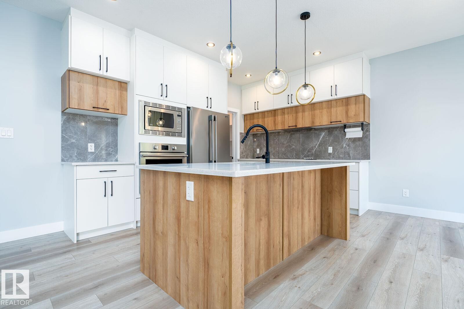 Contemporary kitchen featuring a large central island with a white countertop, wood-finish base, and pendant lighting - 17320 9 Avenue, Edmonton, AB