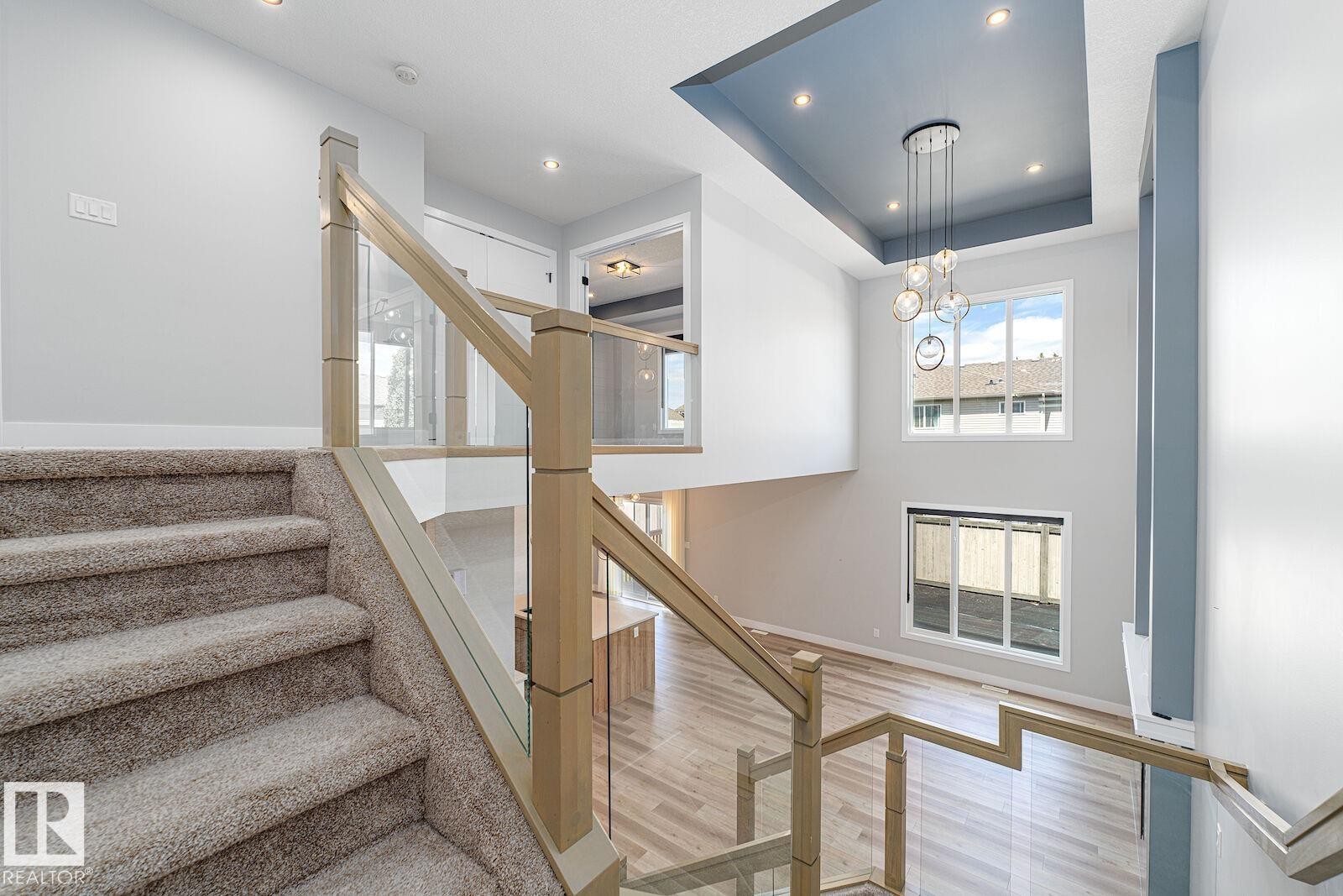 Open concept interior featuring a two-story ceiling, multiple windows, a modern chandelier, light wood-finish flooring, and a staircase with glass panel railing - 17320 9 Avenue, Edmonton, AB