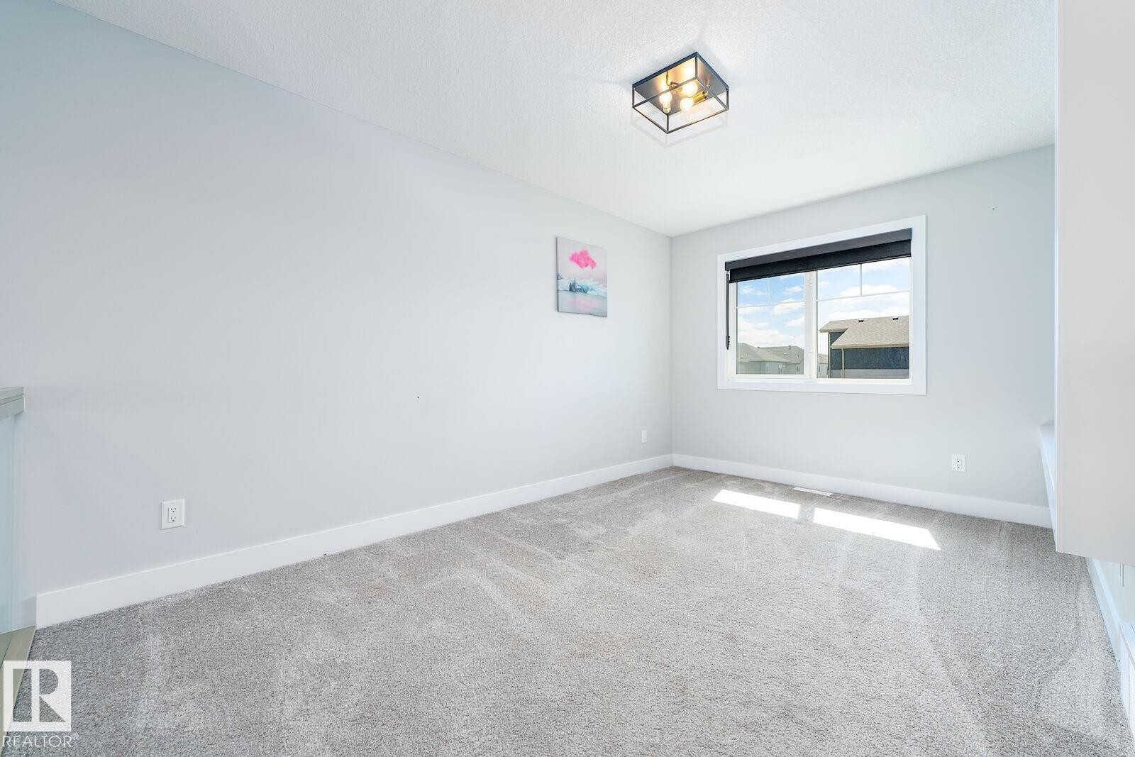Spacious room featuring light gray carpeting, a large window with a dark roller shade, a modern flush-mount light fixture, white baseboards, and light-colored walls - 17320 9 Avenue, Edmonton, AB