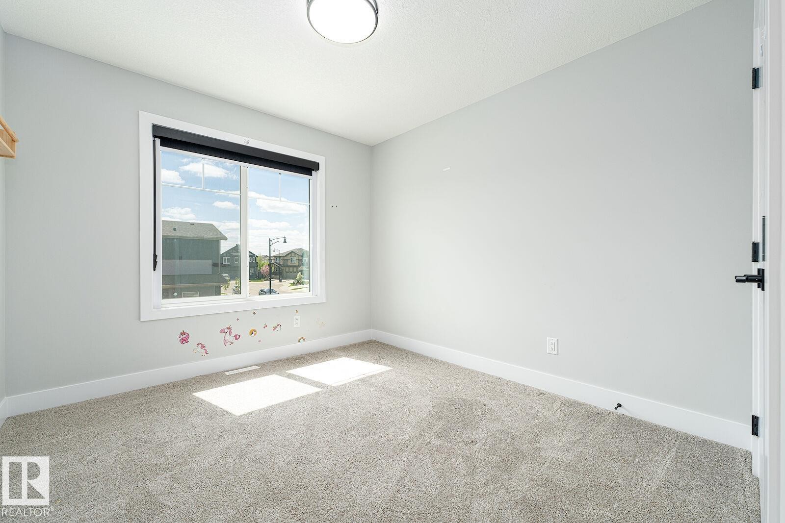 Bright room featuring a large window with black roller blinds, light gray wall paint, white trim, and neutral carpet flooring - 17320 9 Avenue, Edmonton, AB