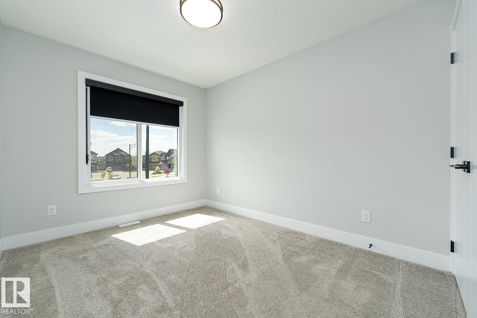 Room with light grey wall paint, plush grey carpet, a white framed window with black roller shade, and a white interior door with matte black hardware - 17320 9 Avenue, Edmonton, AB
