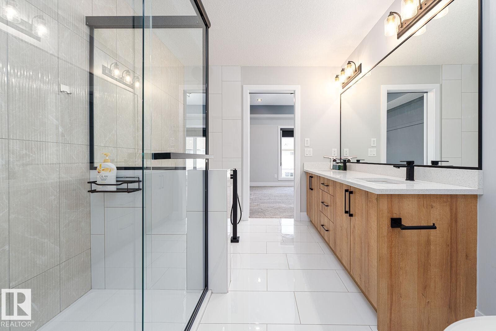 Contemporary bathroom featuring a spacious glass-enclosed shower with light-toned wall tile, a large wood-finish dual vanity with quartz countertops, and matte black hardware - 17320 9 Avenue, Edmonton, AB