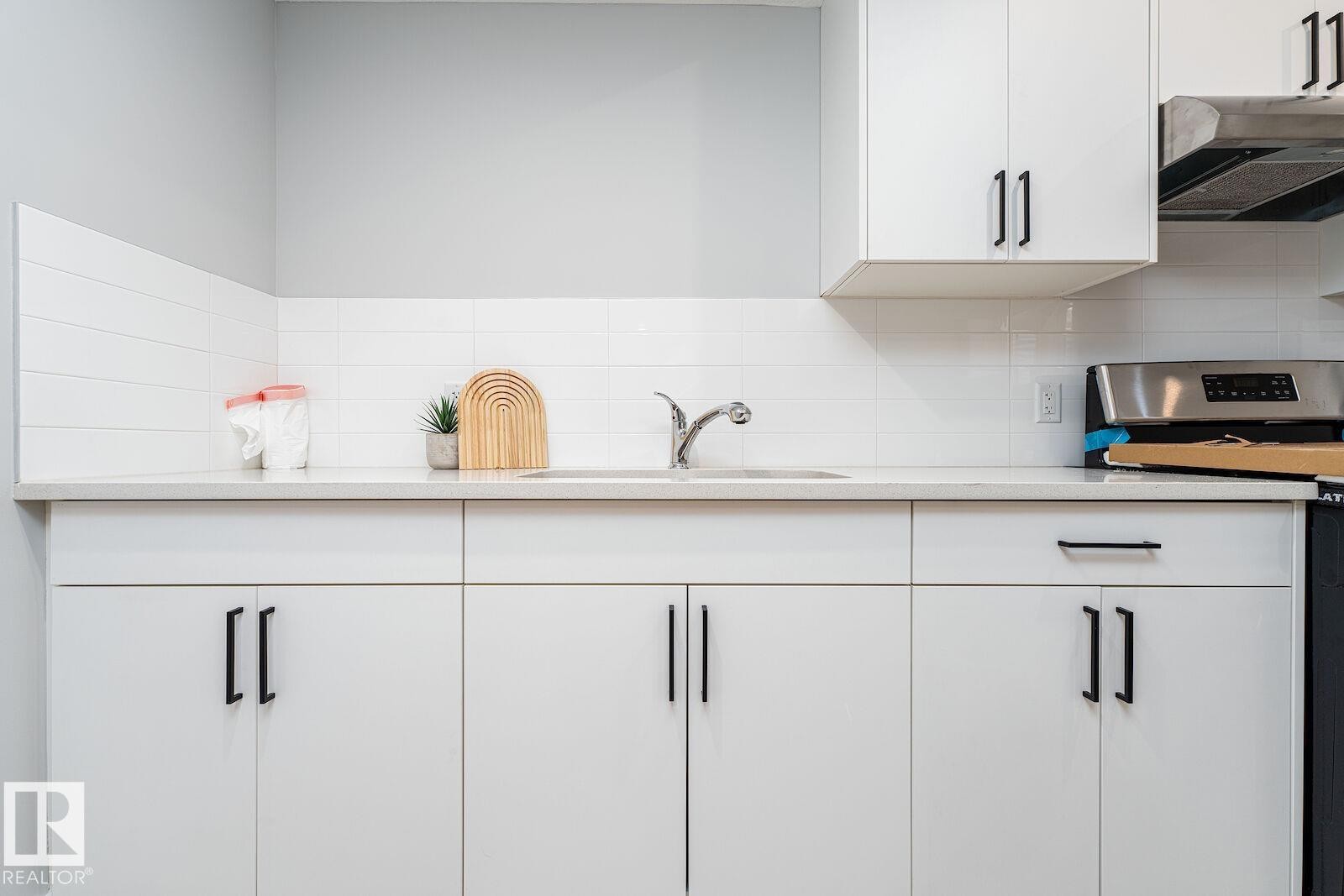 Modern kitchen featuring white cabinetry, light-toned countertops, a stainless steel sink with a chrome faucet, white subway tile backsplash, and stainless steel appliances - 17320 9 Avenue, Edmonton, AB