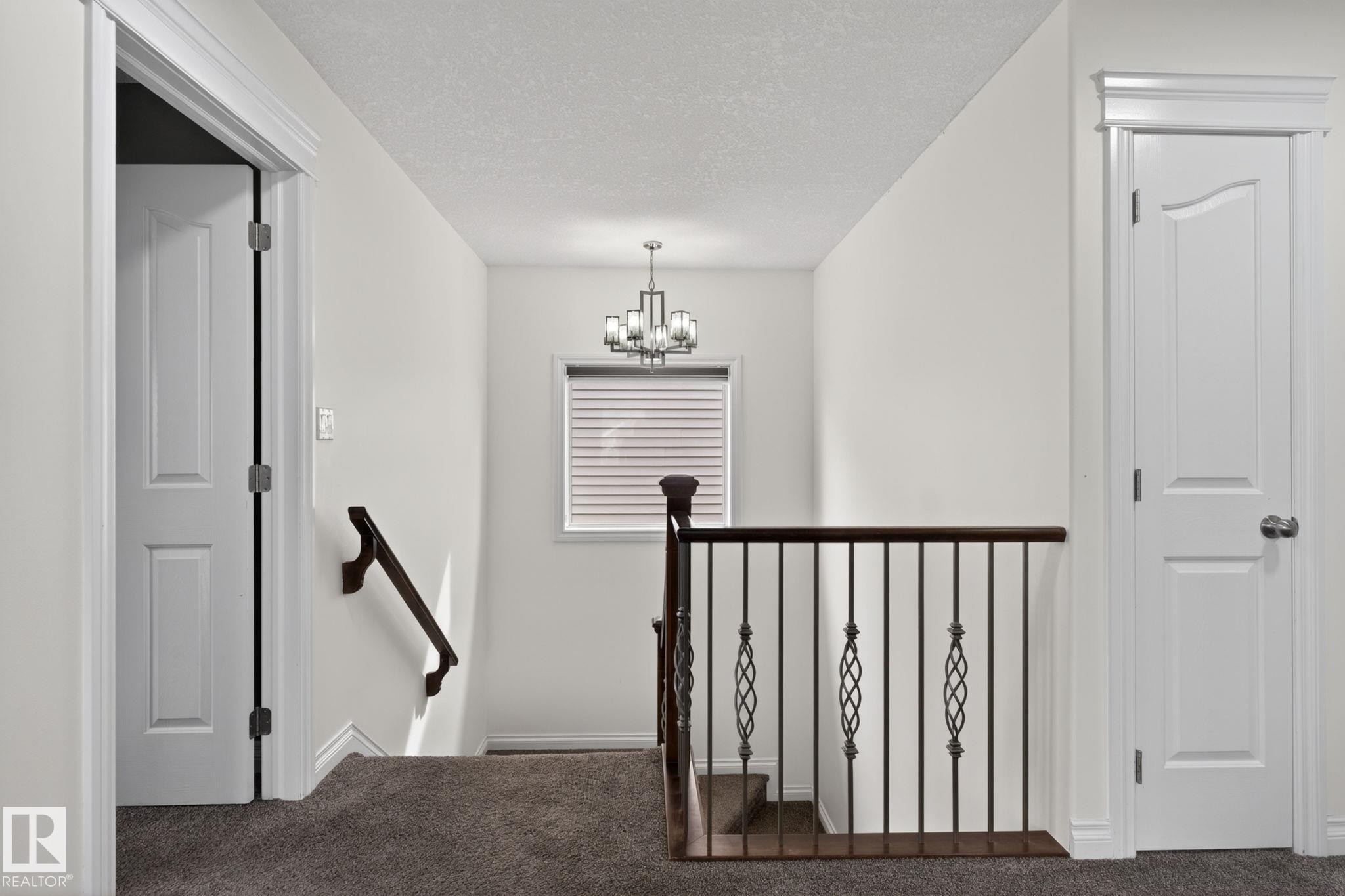Upper-level landing featuring neutral carpeting, a staircase with dark wood railing and wrought iron balusters, a window with horizontal blinds, and a chandelier with a dark finish - 9628 221 Street, Edmonton, AB