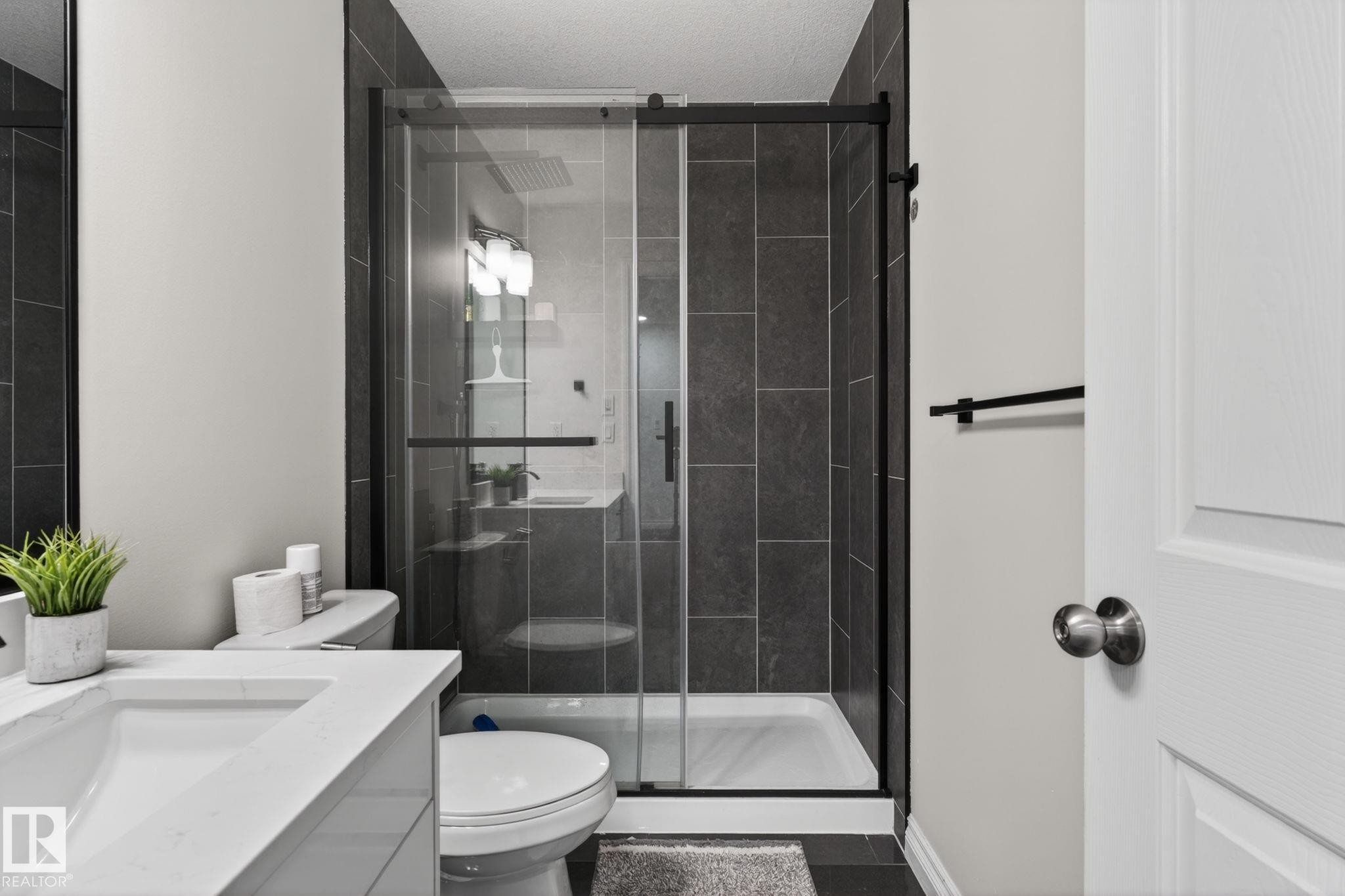 Contemporary bathroom featuring a glass-enclosed shower with dark tile surround, a sleek white toilet, and a vanity with an integrated sink and quartz-finish countertop - 9628 221 Street, Edmonton, AB