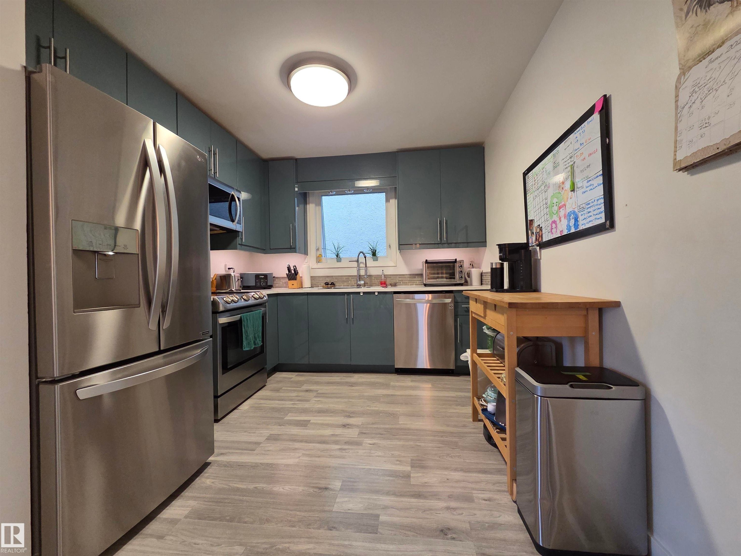 Galley kitchen featuring full-height teal cabinetry, stainless steel appliances, light-toned countertops, and wood-finish flooring - 2514 149 Avenue, Edmonton, AB