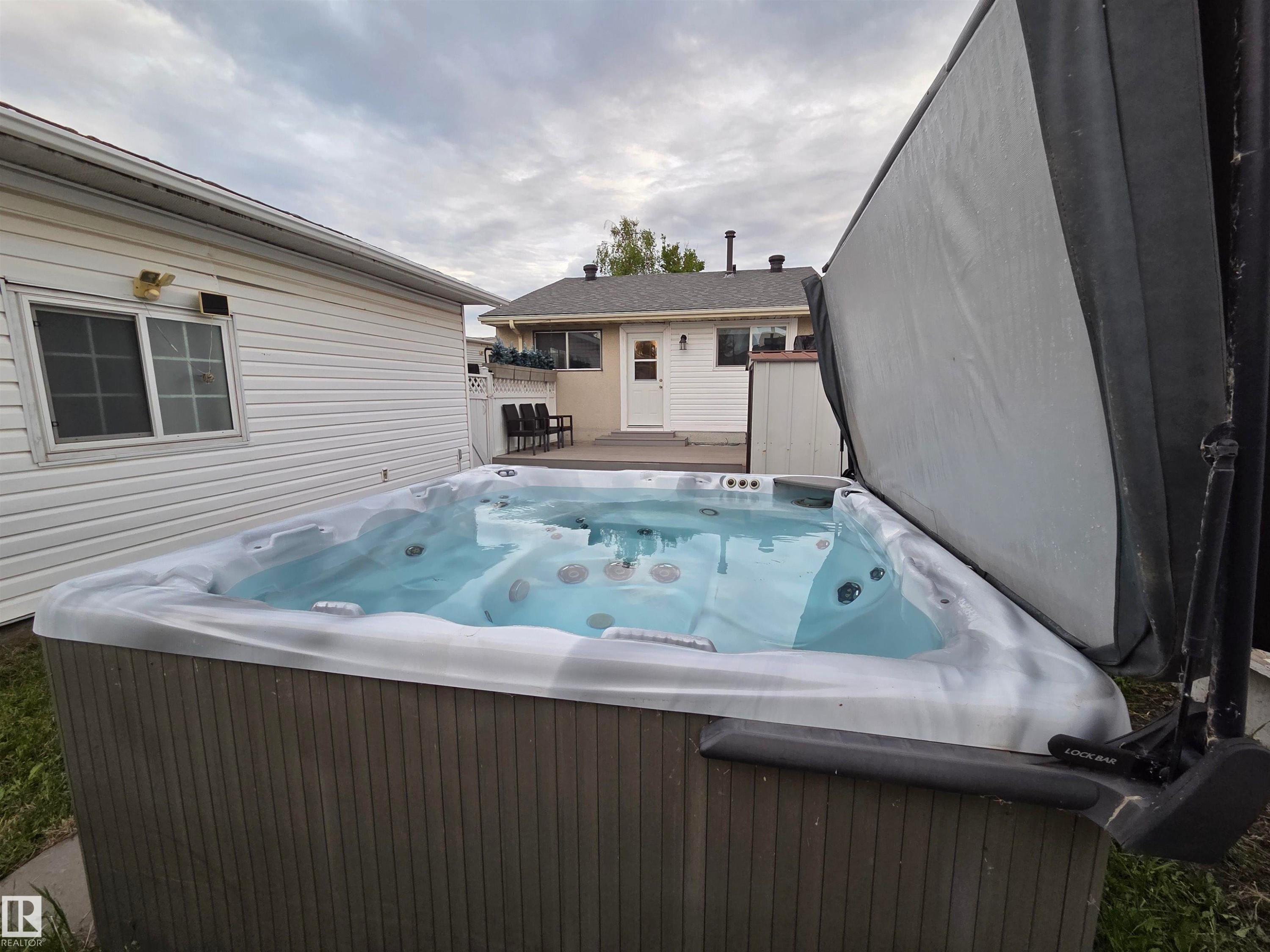 Exterior spa with a partially opened cover, a white vinyl-sided building, and a rear deck with a white door - 2514 149 Avenue, Edmonton, AB