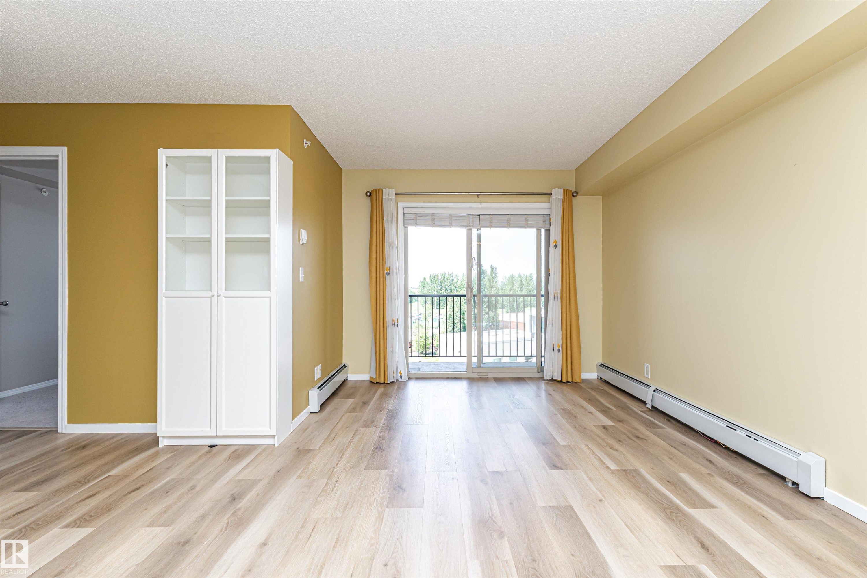 Spacious living area featuring wood-finish flooring, a built-in white cabinet with glass-front uppers, and a sliding glass door leading to a balcony - 404 12045 22 Avenue, Edmonton, AB