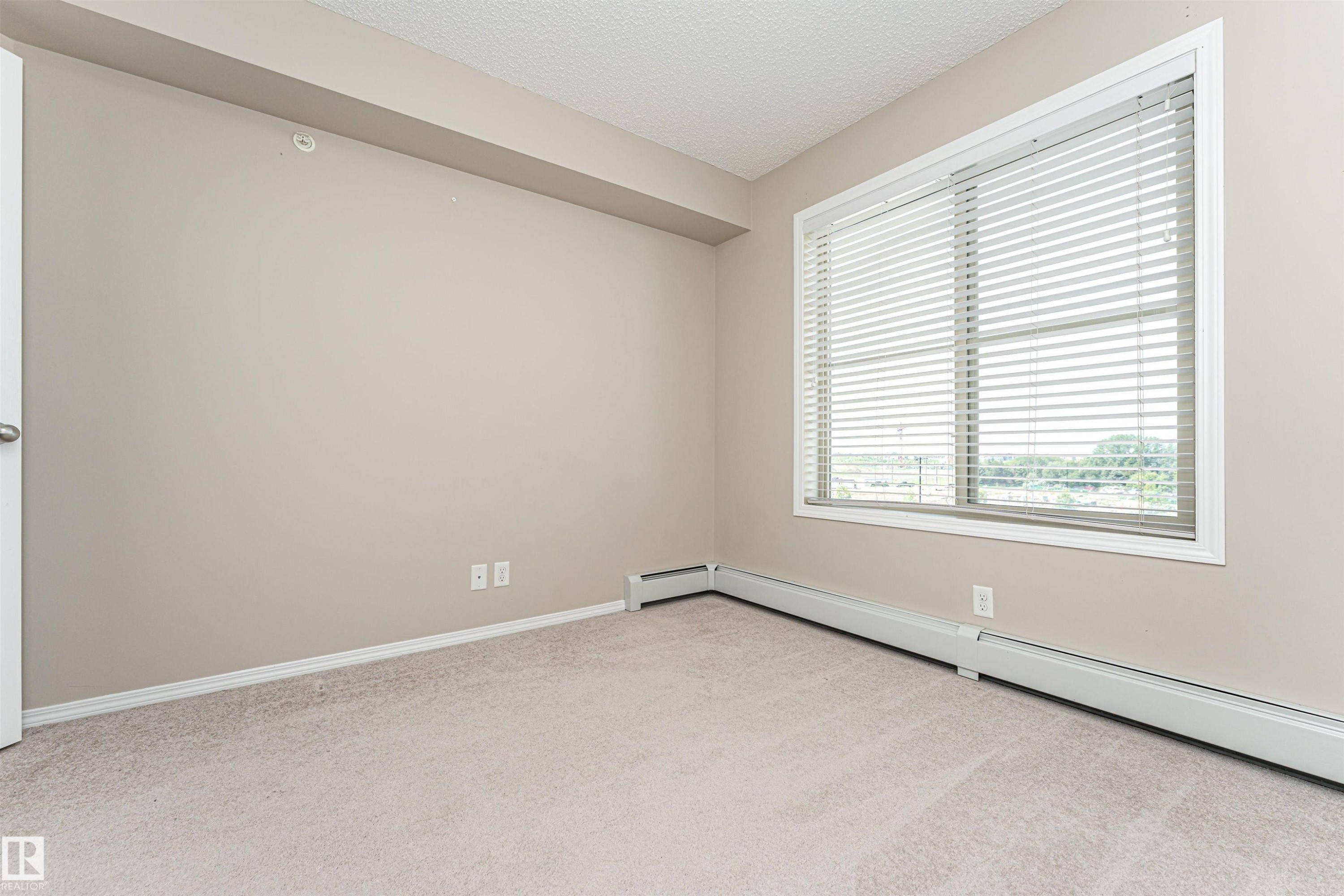 Carpeted interior room featuring neutral wall paint, white trim, baseboard heating, and a large window with horizontal blinds - 404 12045 22 Avenue, Edmonton, AB