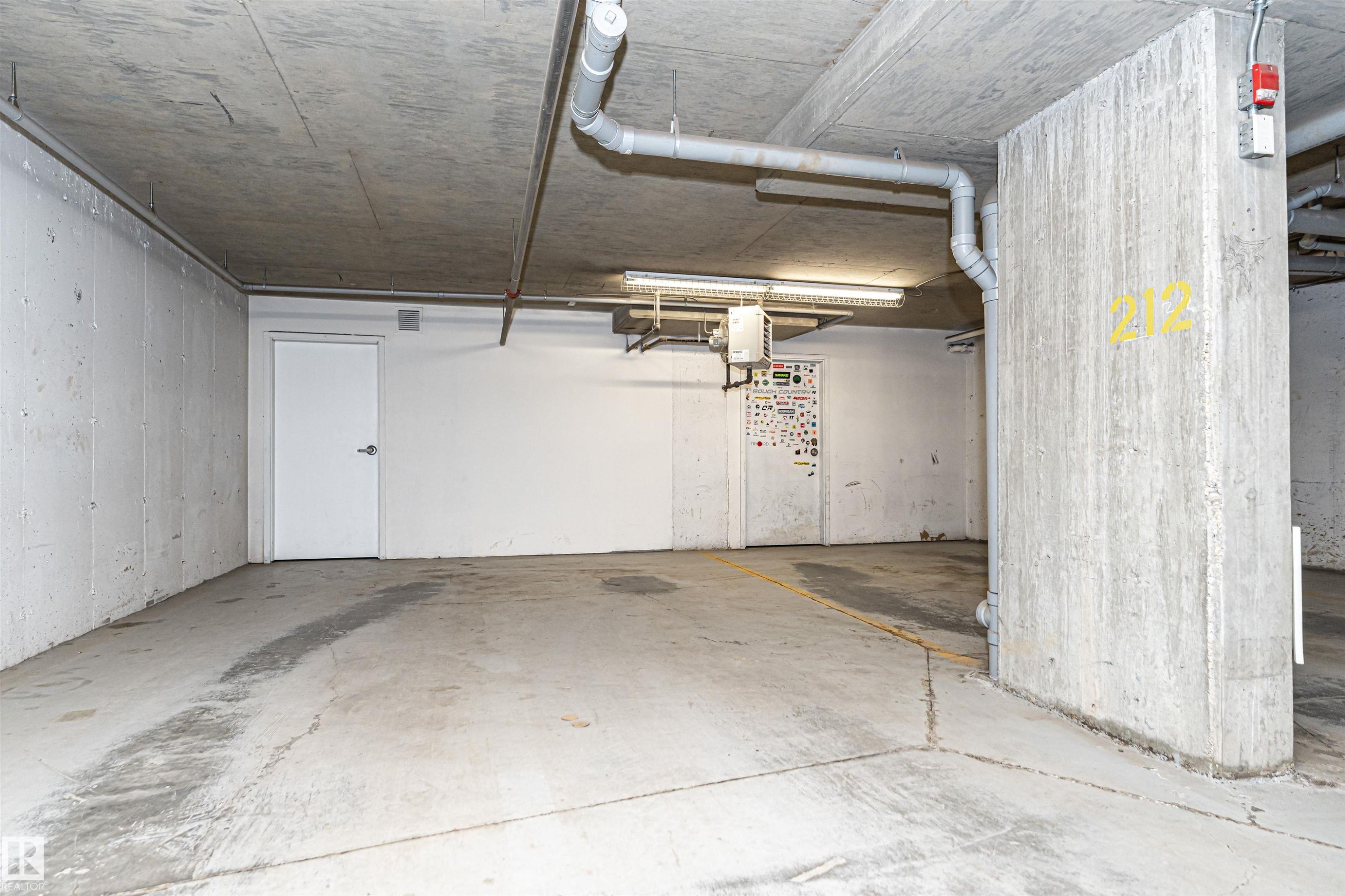Concrete parking space featuring exposed ceiling pipes, fluorescent lighting, and a white access door - 404 12045 22 Avenue, Edmonton, AB