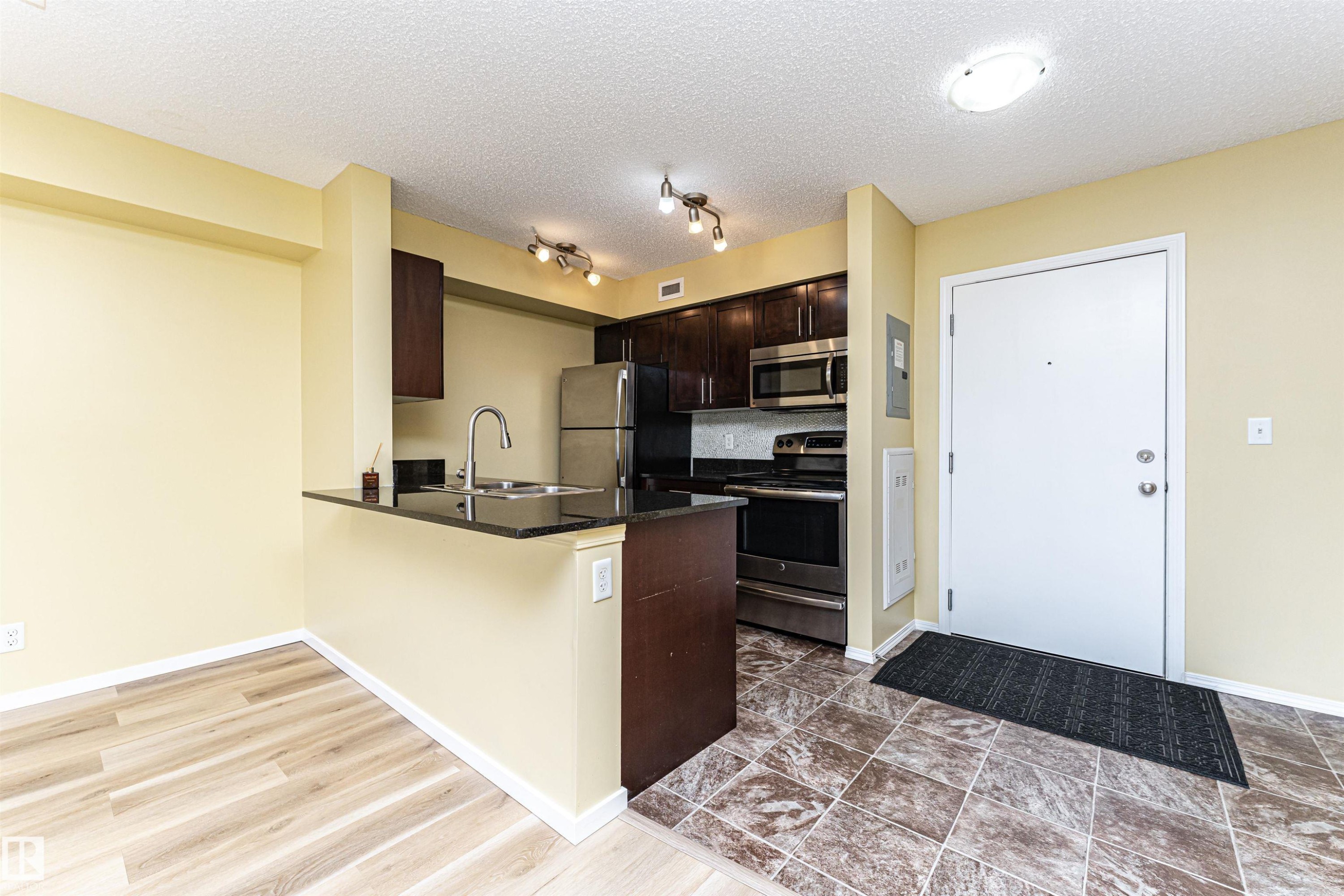 Compact kitchen featuring dark wood cabinetry, stainless steel appliances, black countertops, and a white subway tile backsplash - 404 12045 22 Avenue, Edmonton, AB