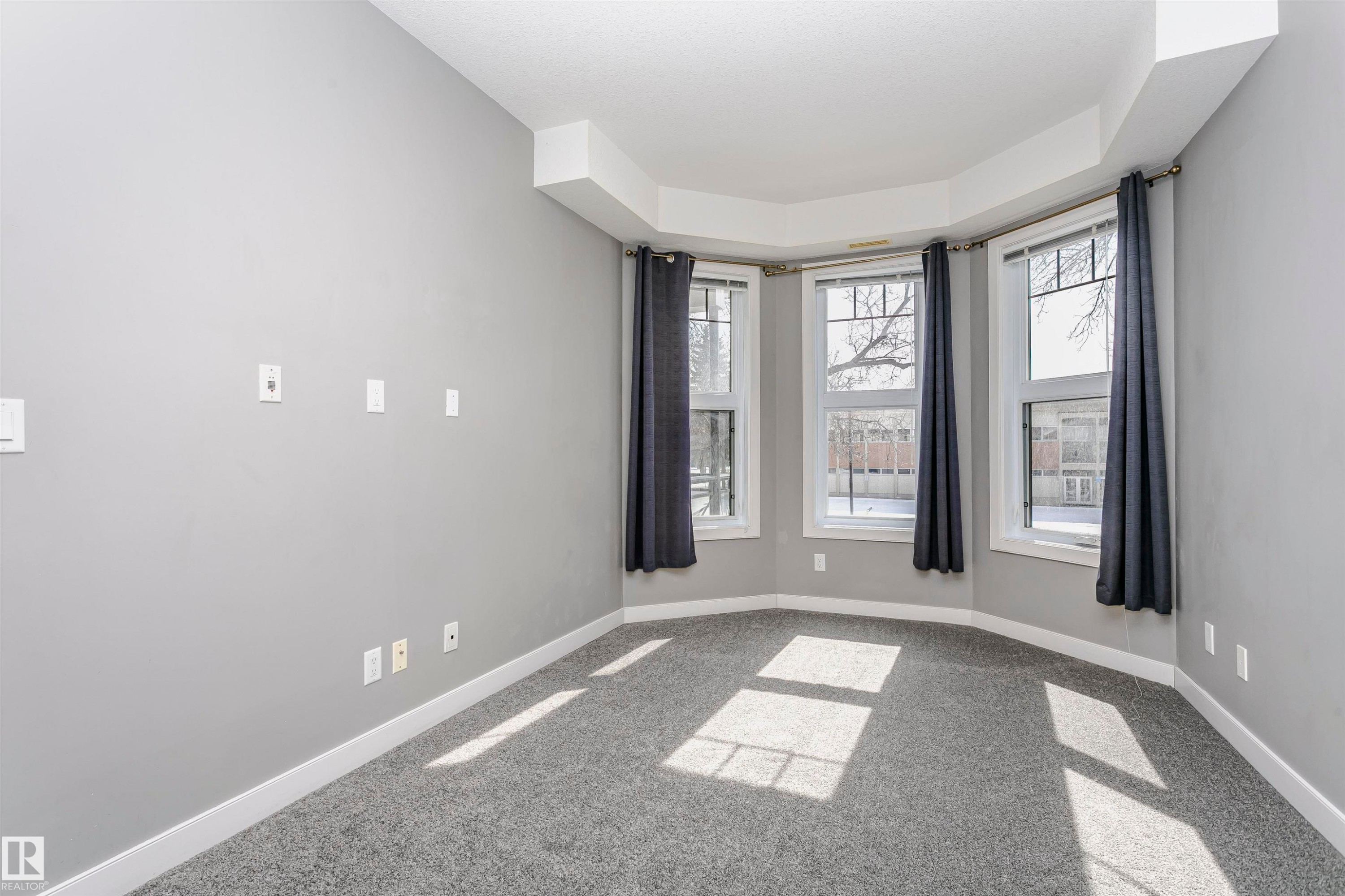 Bay window with three individual windows, light gray walls, dark gray carpeting, and white baseboards - 211 10808 71 Avenue, Edmonton, AB