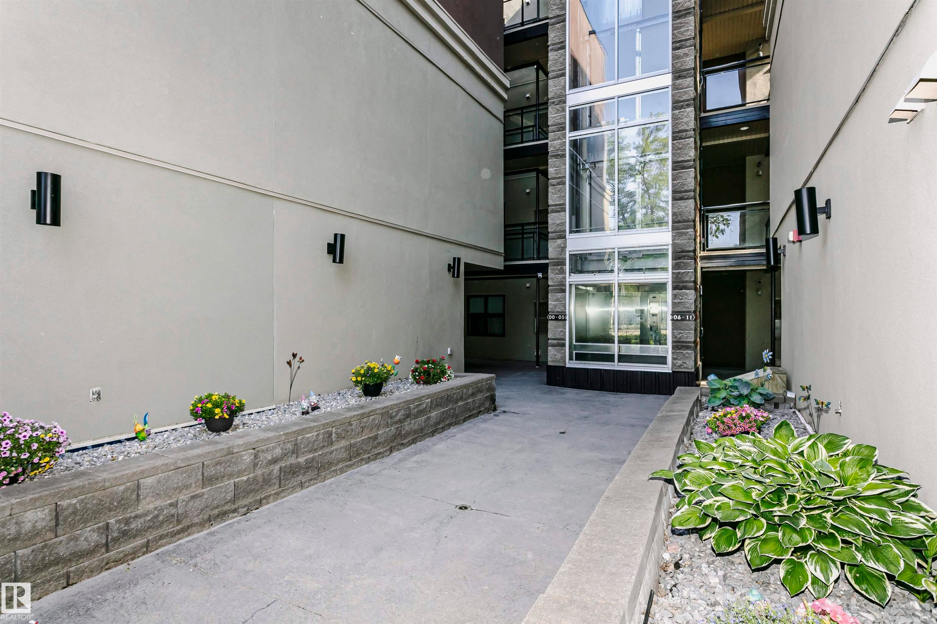 Exterior walkway featuring concrete paving, stone retaining walls, and a multi-story glass elevator shaft with stone detailing - 211 10808 71 Avenue, Edmonton, AB