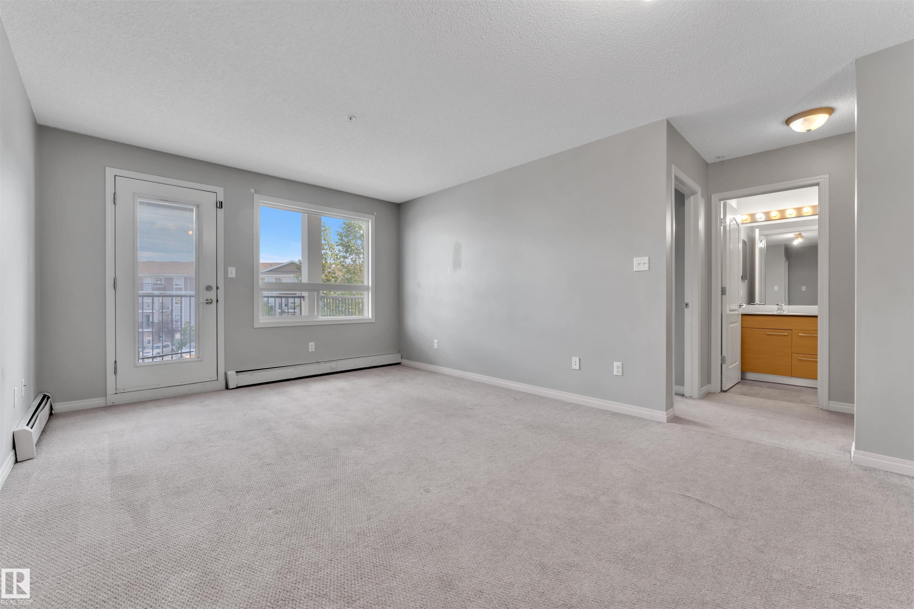 Spacious living area featuring light gray carpeting, neutral wall tones, and a glass-paneled door leading to a balcony - #3302 9351 Simpson Dr Nw, Edmonton, AB