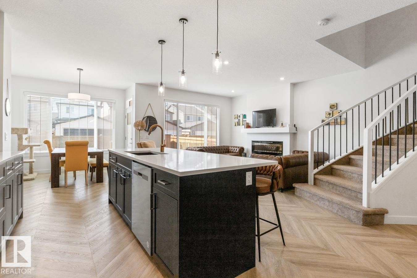 Open-concept living space featuring wood-finish flooring with a herringbone pattern, a kitchen island with a white countertop, a built-in electric fireplace, and a staircase with a black metal spindle railing - 117 Paisley Gate Gate, Edmonton, AB