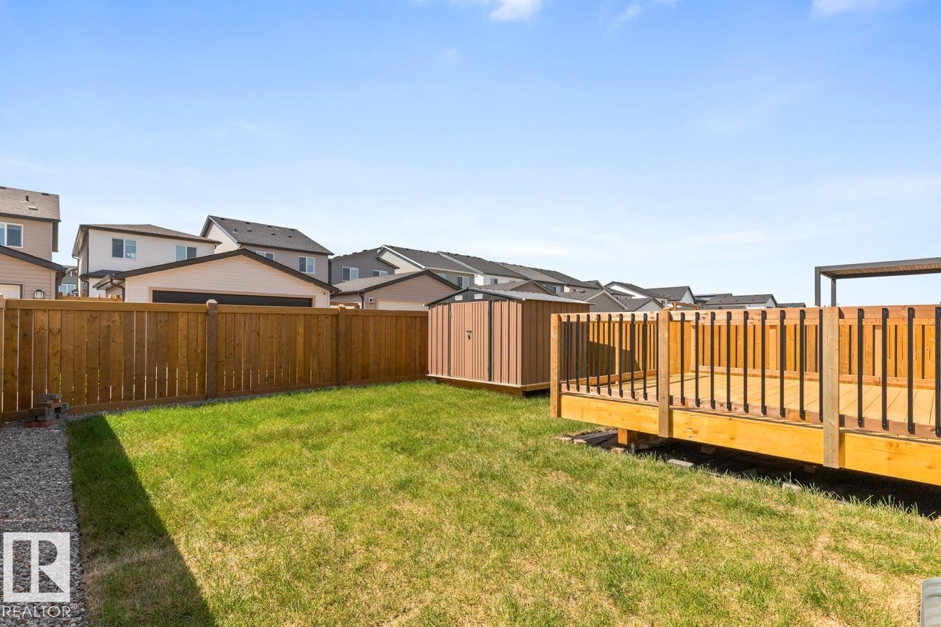 Expansive backyard featuring a natural wood deck with black metal balusters, a gravel border, lush green grass, a natural wood privacy fence, and a storage shed - 117 Paisley Gate Gate, Edmonton, AB