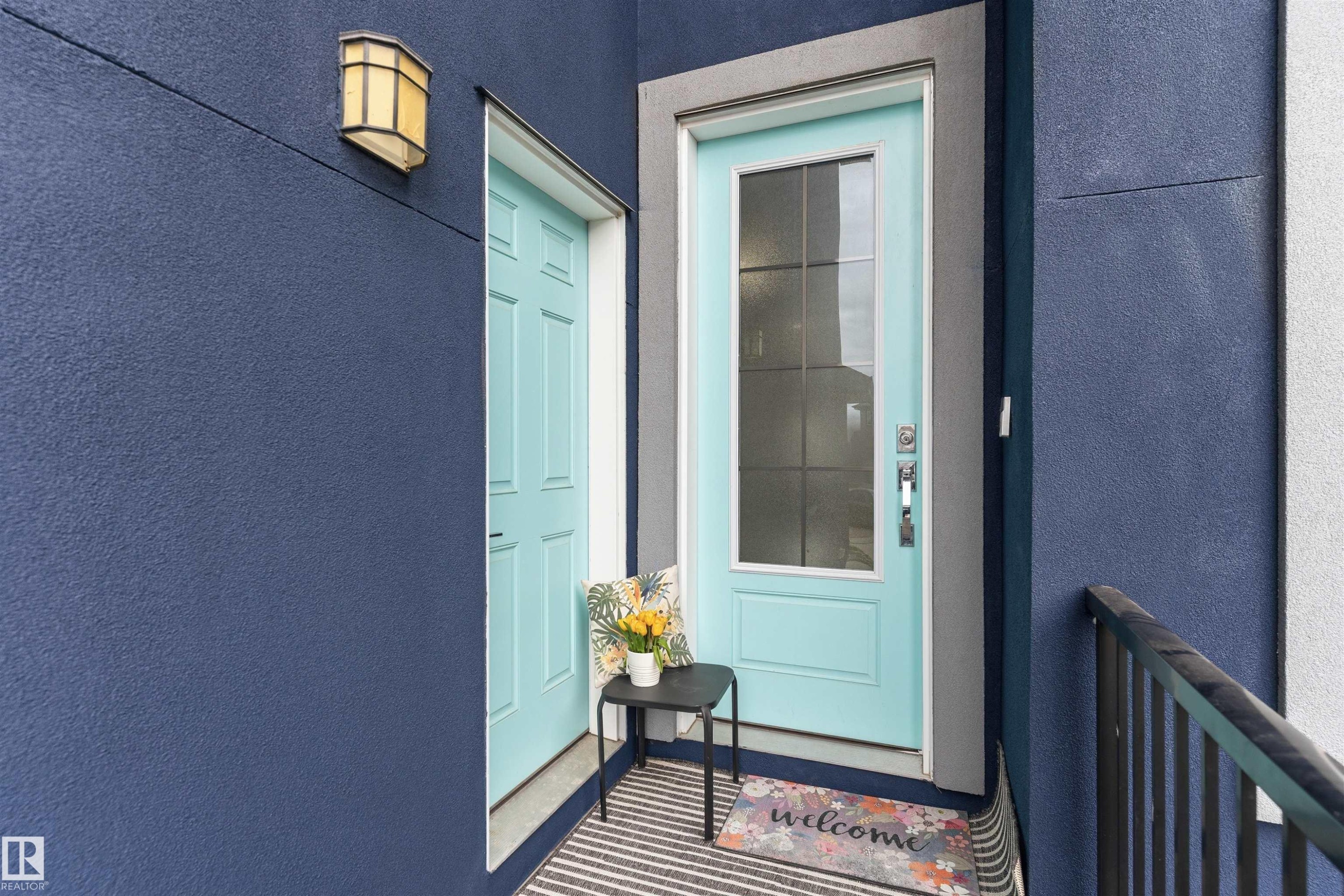 Contemporary exterior entrance featuring a light blue glass-paneled door with brushed nickel hardware, a coordinating solid light blue door, textured dark blue stucco siding, and a black metal railing - 3312 11 Avenue, Edmonton, AB