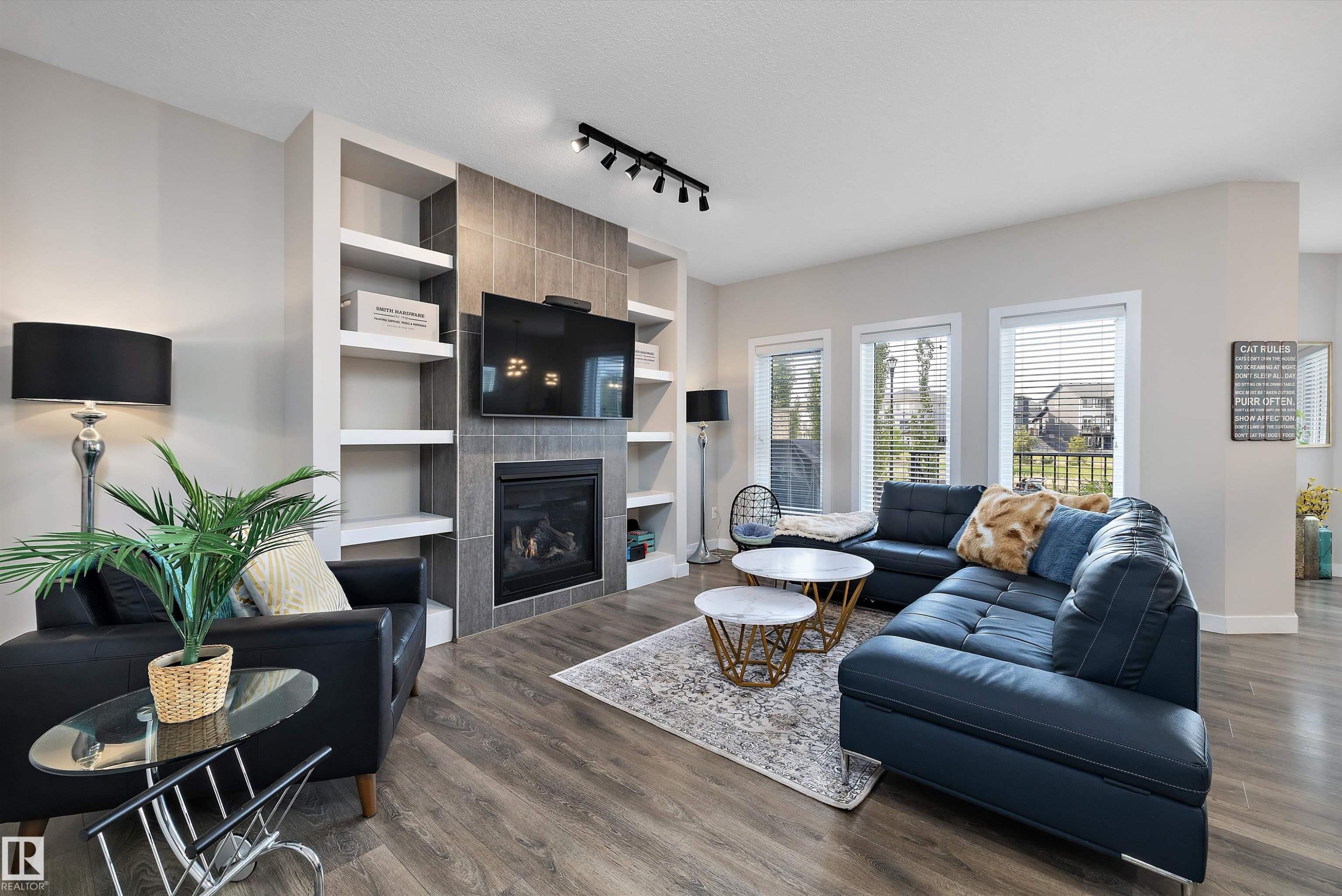 Living area featuring wood-finish flooring, a tiled gas fireplace, built-in shelving, track lighting, and multiple windows with blinds - 363 Cy Becker Boulevard, Edmonton, AB - Indoor Photo Showing Living Room With Fireplace